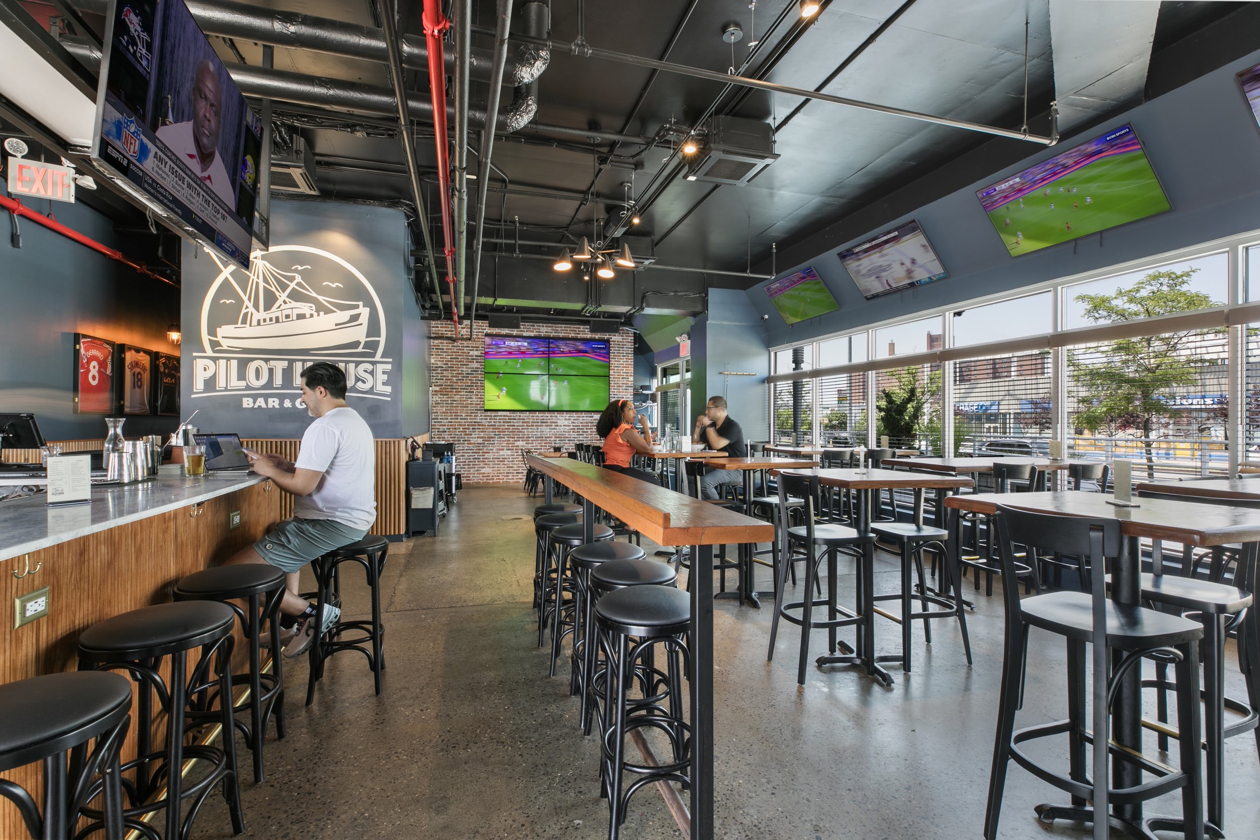 Interior of a sports bar with multiple TV screens showing a football game, high tables with stools, and a few patrons seated and dining. There is a large sign on the wall that reads 'PILOT HOUSE BAR & GRILL,' and the decor features exposed pipes and 