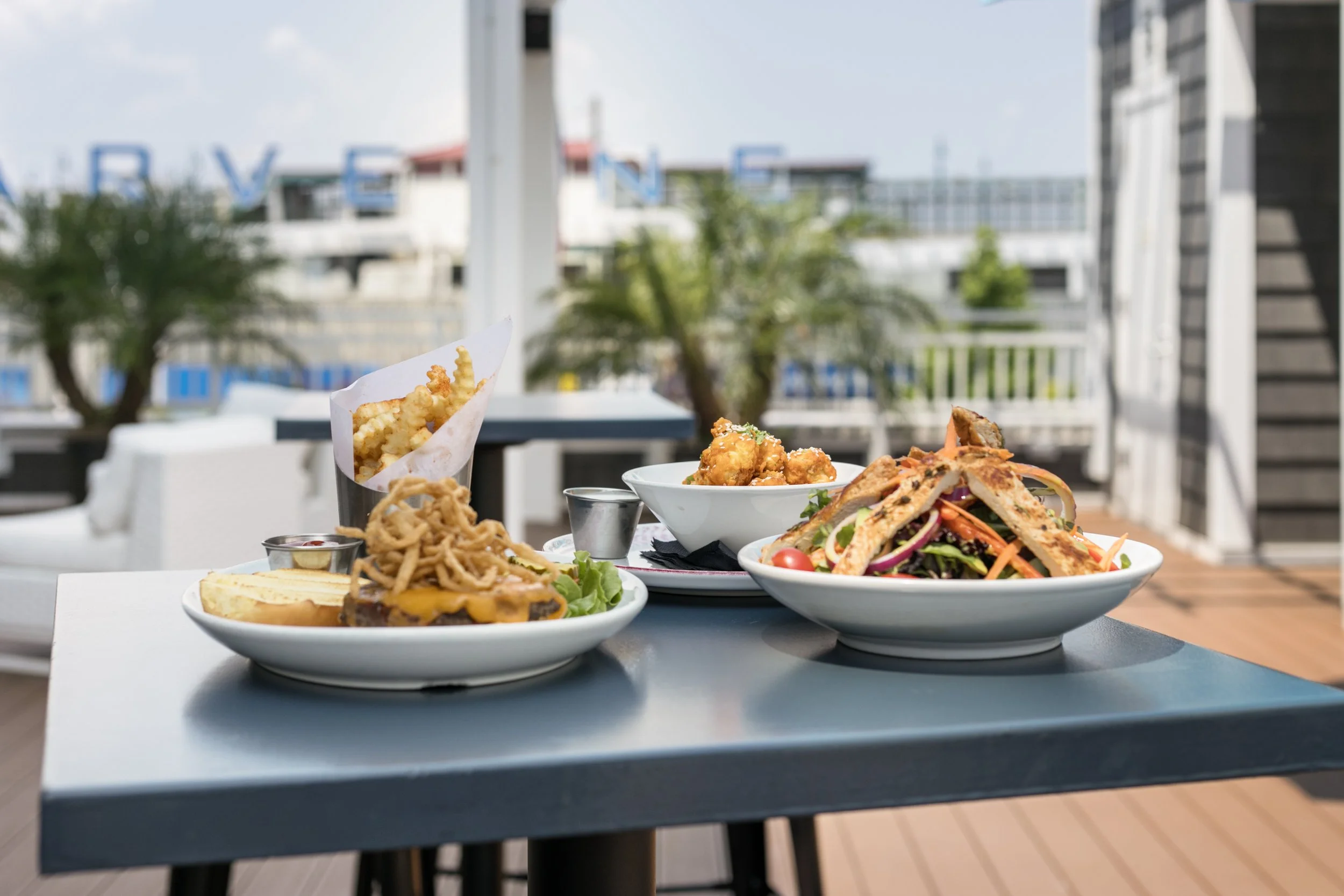Several plates of food, including a burger, fried chicken, salad, fries, and a wrap, are on a table outdoors with blurred buildings, trees, and a balcony in the background.