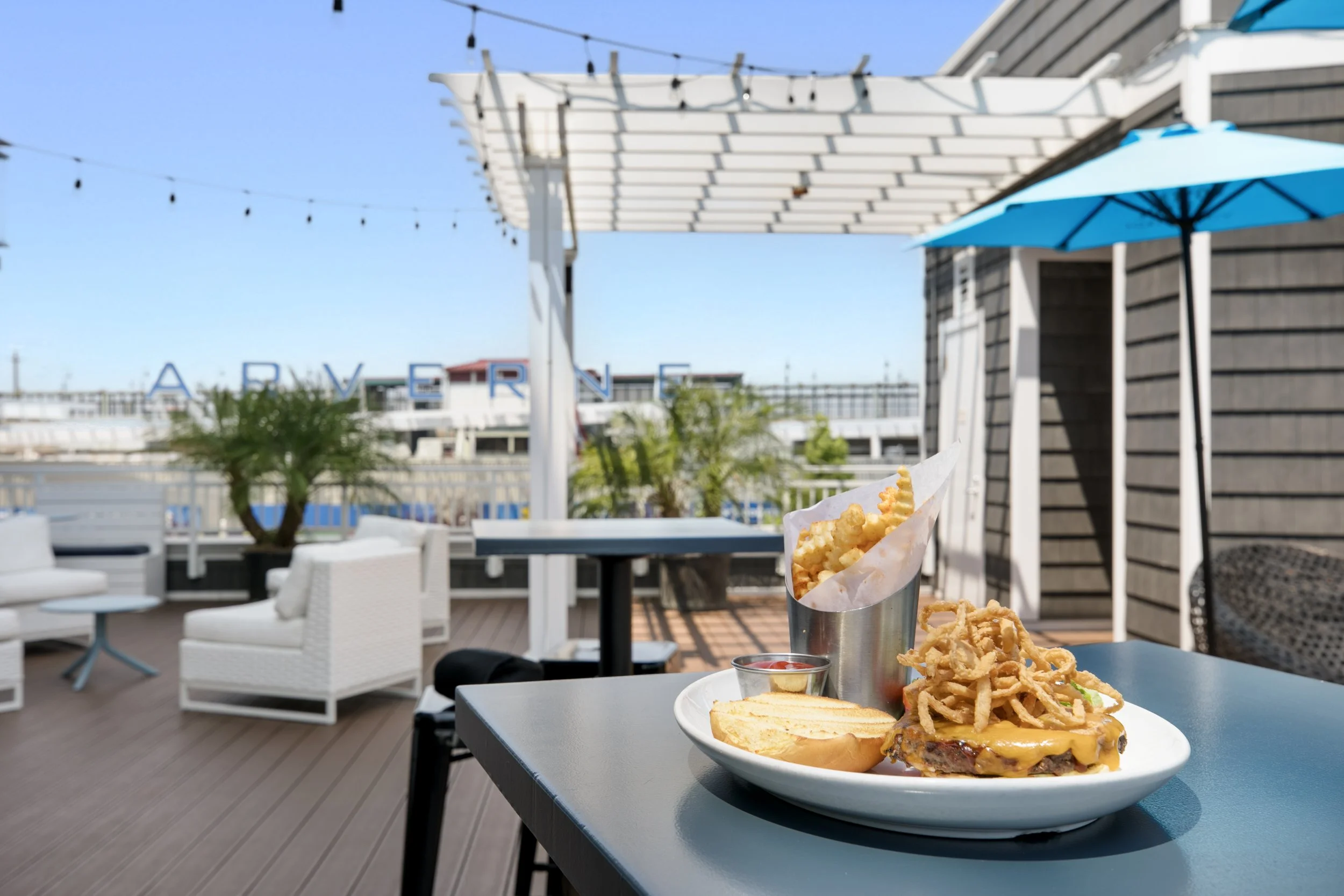 Outdoor dining scene with a plate of cheeseburger, fries, and onion rings on a dark table, with patio furniture and string lights under a blue sky.
