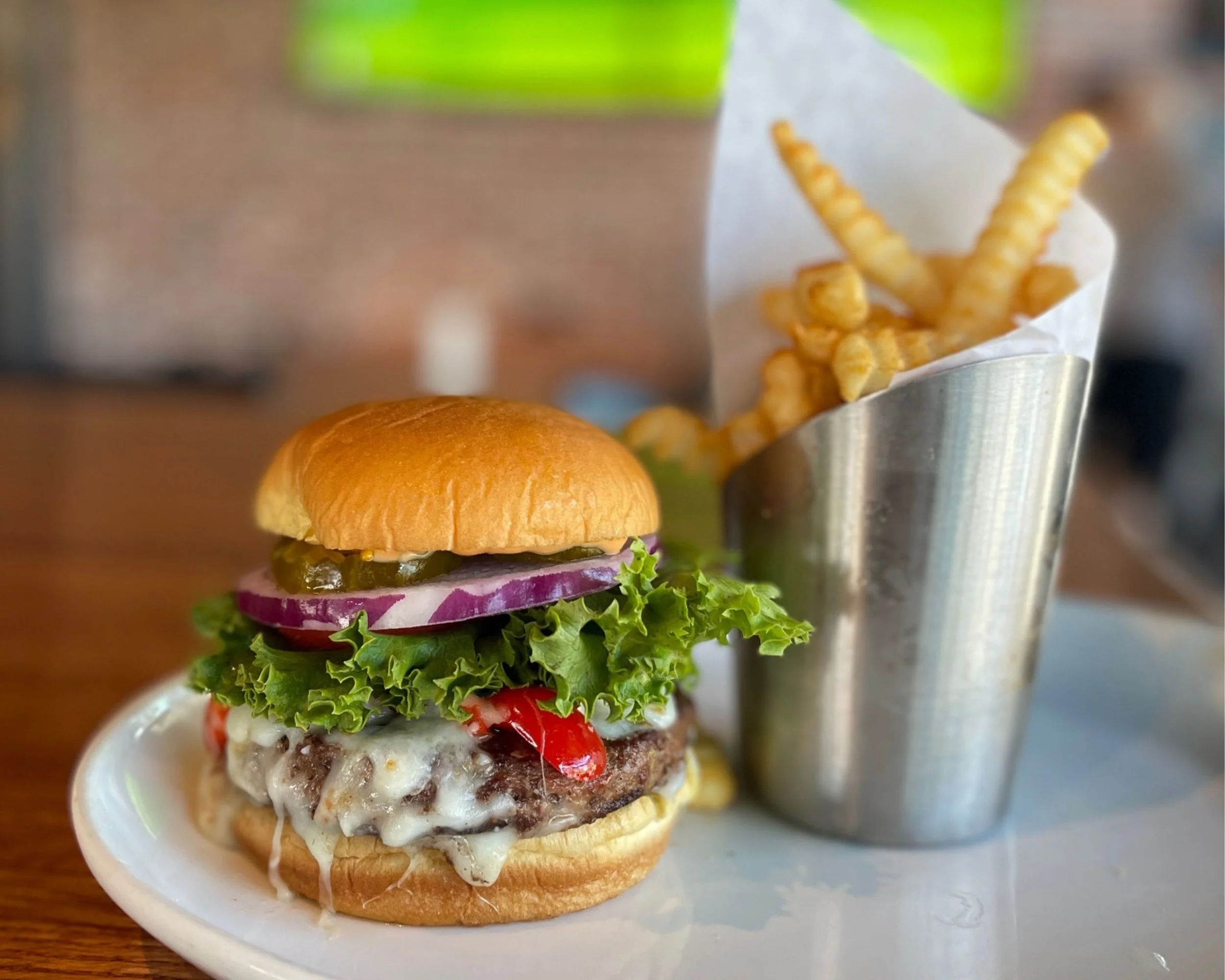 Close-up of a hamburger with lettuce, tomato, onion, melted cheese, and a beef patty, served in a bun, placed on a white dish. In the background, a serving of crinkle-cut French fries in a metal container.