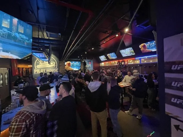 Crowd gathered inside a sports bar with multiple screens displaying UFC events, and a large sign reading 'Pilot House' on the wall.