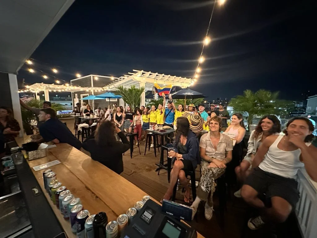 People gathered on a rooftop at night, some watching a sporting event, with string lights overhead, a bar counter on the left, and a view of the city in the background.