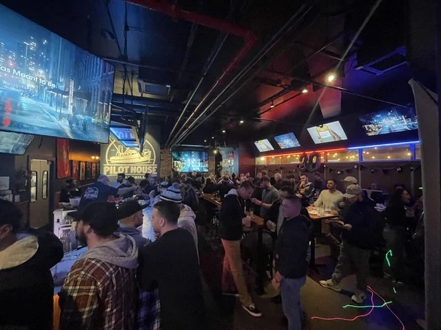 Crowd of people socializing and playing arcade games inside a bar with multiple screens, neon lights, and a sign reading 'PILOT HOUSE' in the background.