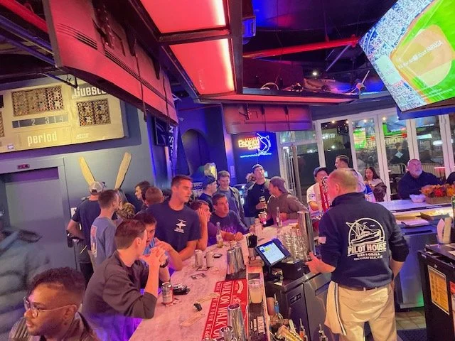 People gathered at a bar with illuminated screens, a large TV showing a sports game, and a chalkboard scoreboard display.