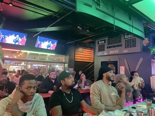 People sitting at a bar or restaurant, watching a sports game on multiple TV screens, with an illuminated American flag on the wall and a game score displayed.