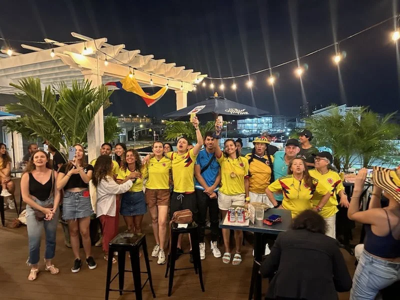 Group of people gathered at an outdoor event at night, some wearing Colombian soccer jerseys, celebrating and cheering under string lights. There are palm trees, umbrellas, and a decorative bandera (flag) in the background.