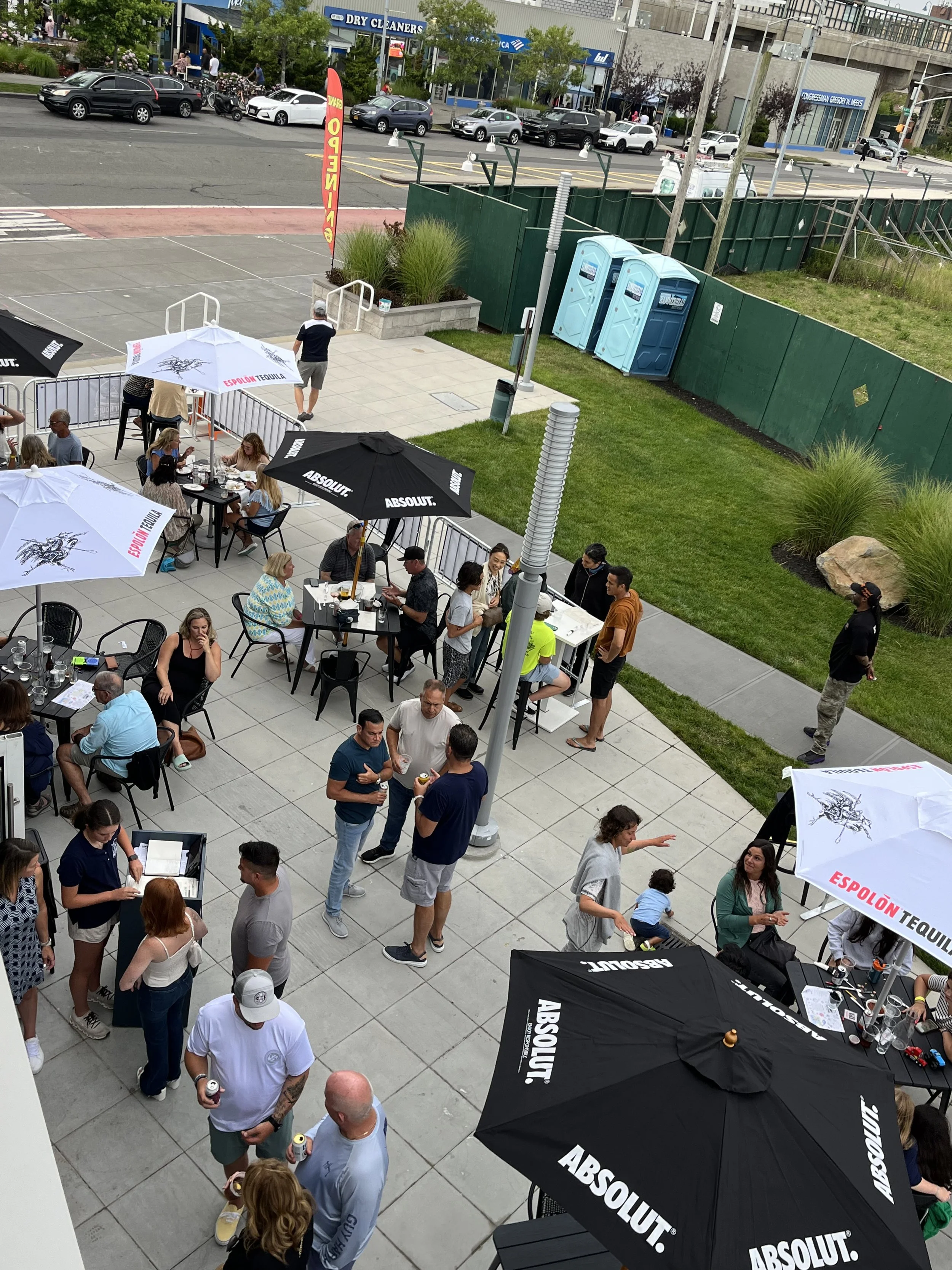 Crowd of people gathering outside at a food or drink event with tables, chairs, and umbrellas branded with 'Absolut' and 'Espolòn Tequila'. Some are standing in line, chatting, or using their phones. The event is set on a sidewalk with a grassy area,