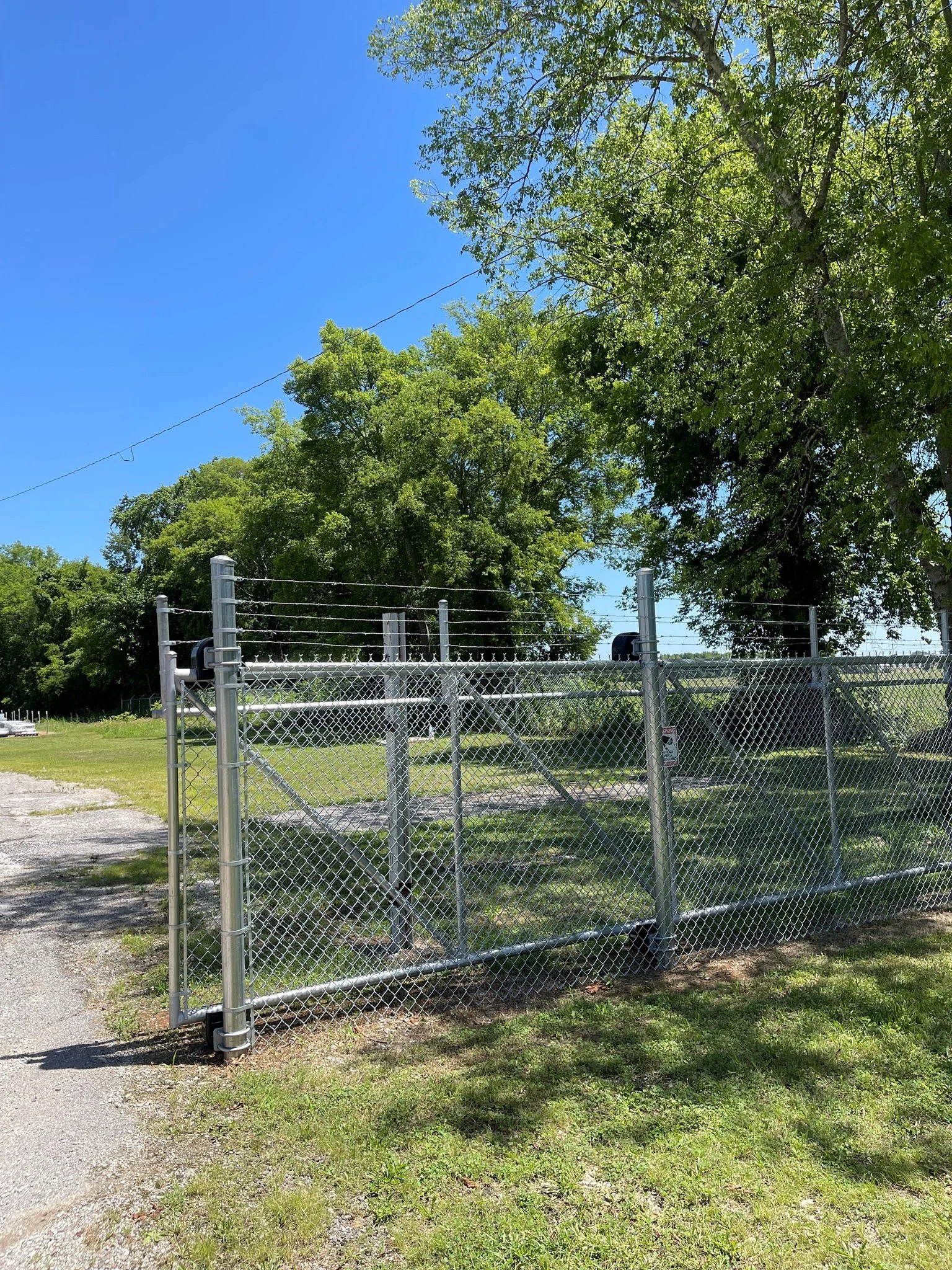 A galvanized, commercial chain-link fence with a cantilever gate, set in a grassy area with trees and a clear blue sky in the background.