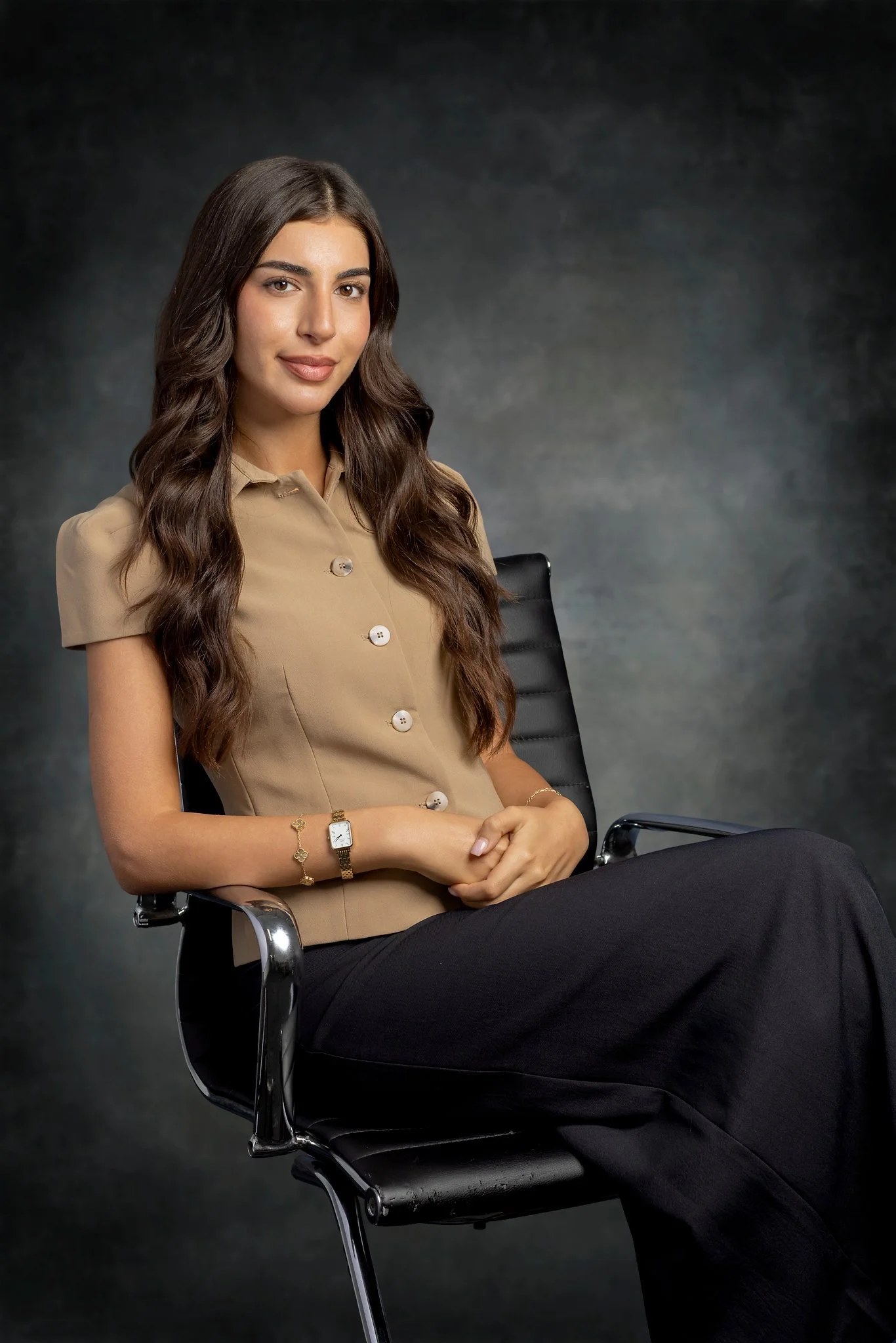 A young woman with long, wavy brown hair, sitting in a black leather office chair against a dark, textured background. She is wearing a beige button-up blouse with short sleeves, black pants, a wristwatch, and a bracelet, and looking confidently at the camera.