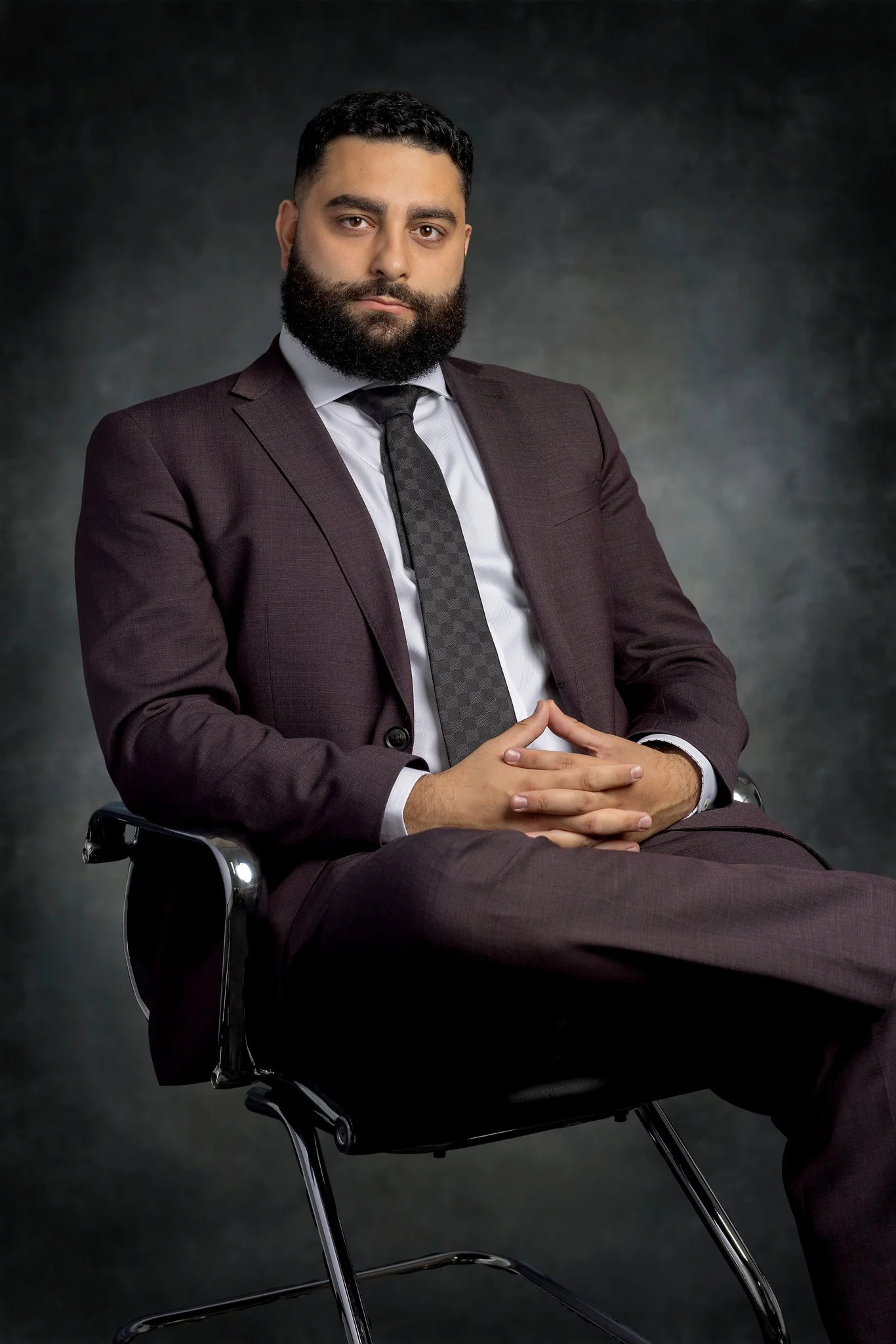 A man with a beard and short hair sitting in a black chair, wearing a dark suit and tie, with hands clasped on his lap, against a dark textured background.