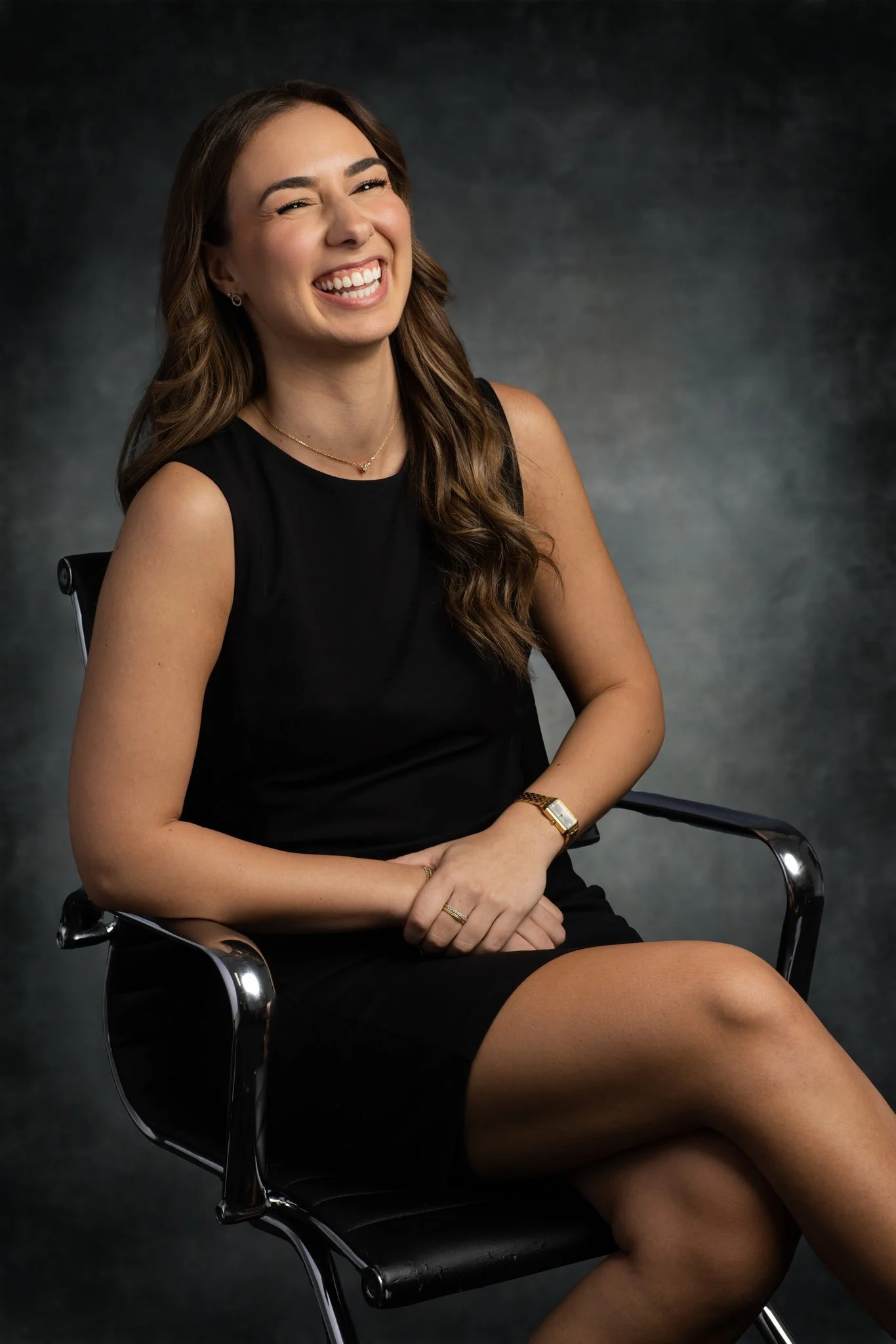 A woman sitting on a black office chair, wearing a sleeveless black dress, smiling and laughing, with a dark textured background.