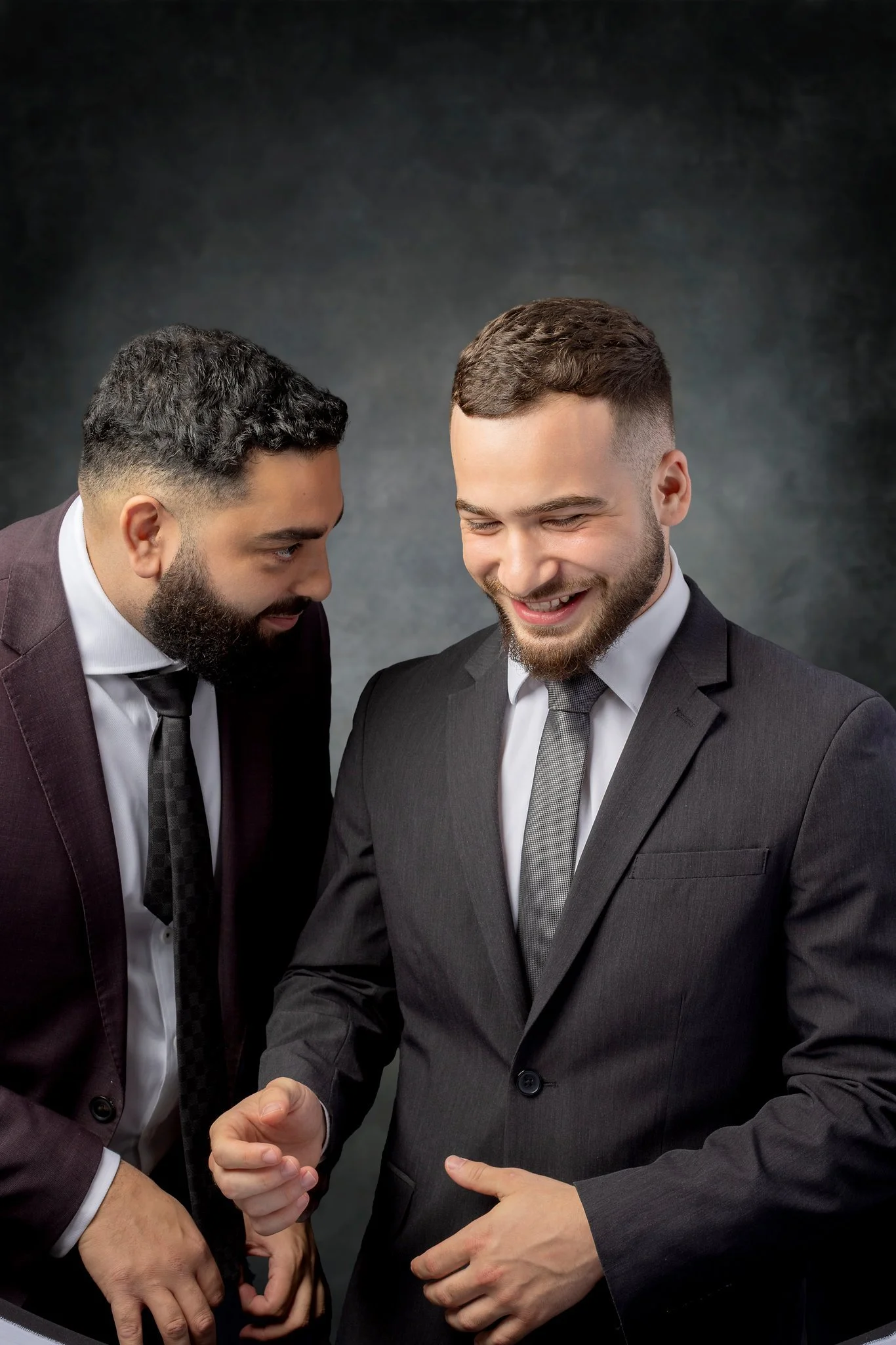 Two men in suits smiling and whispering to each other, standing against a dark gray background.