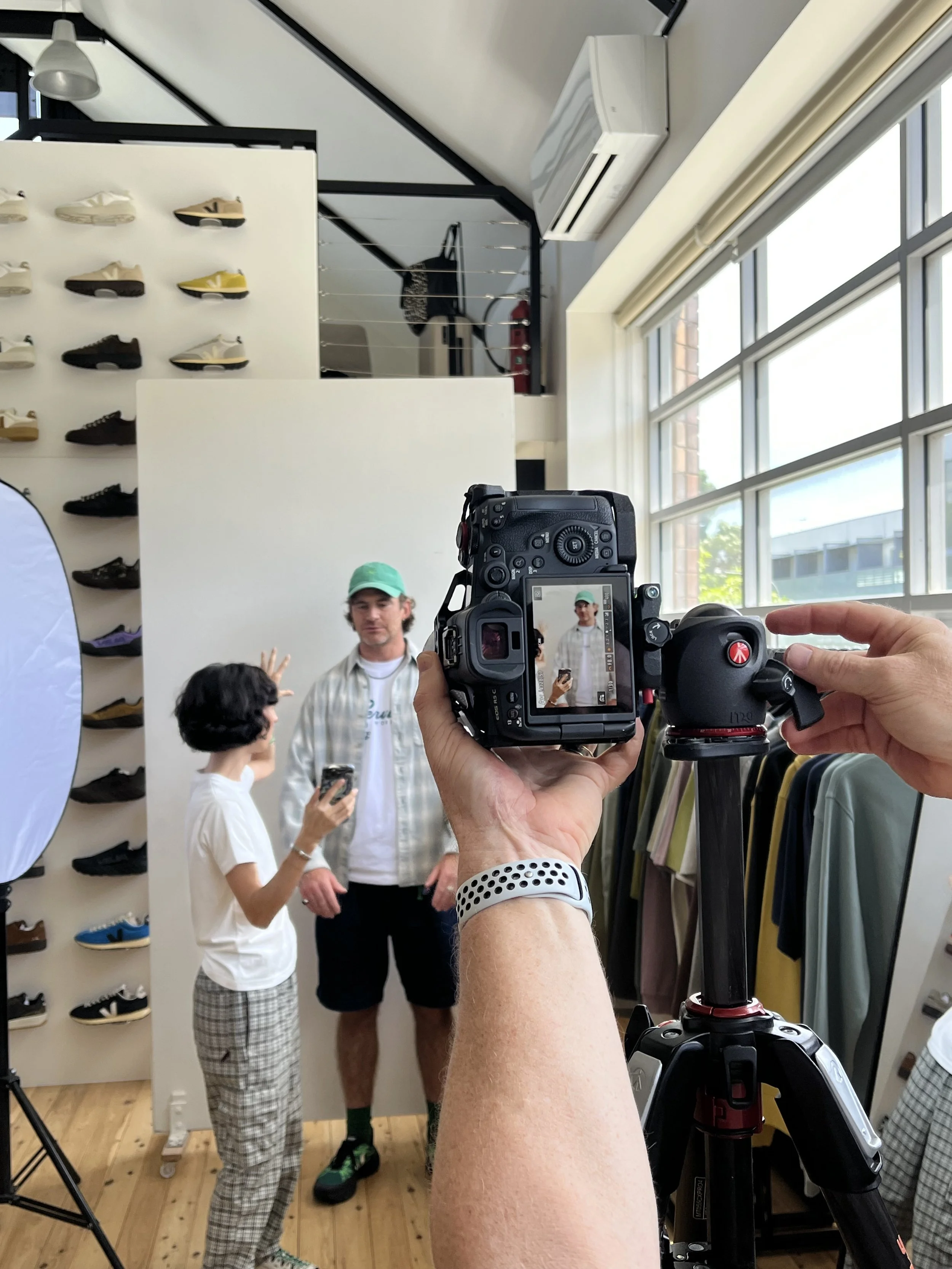 Two people being filmed in a shoe store, with wall display of various sneakers and large windows letting in natural light.