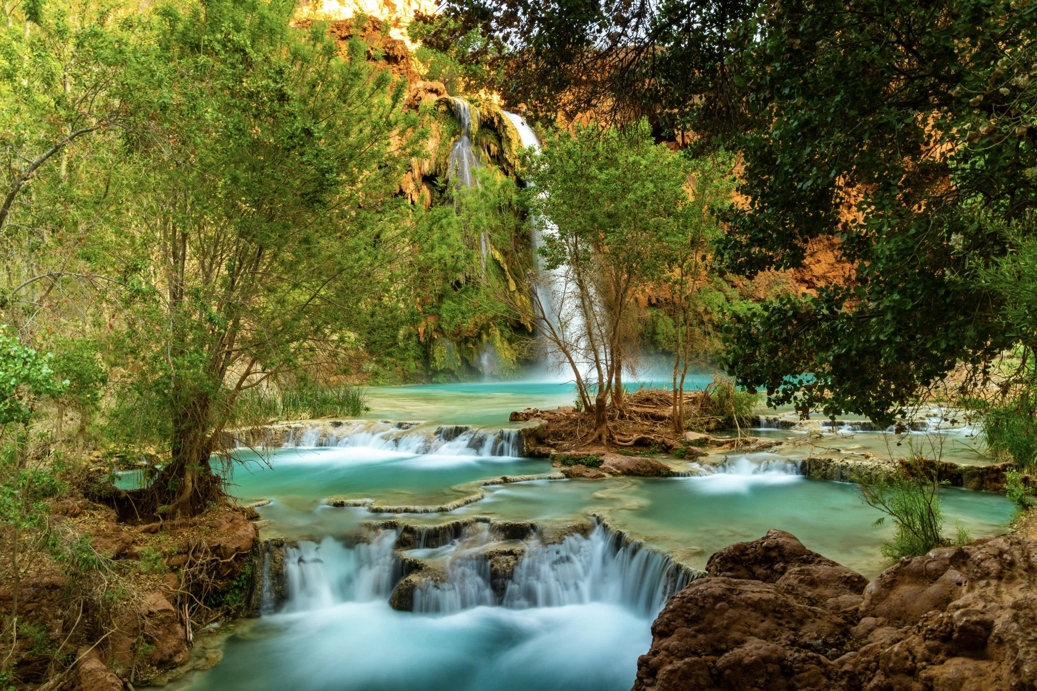 A lush, turquoise cascade of waterfalls and natural pools beneath Havasu Falls in the Grand Canyon, surrounded by green trees and red canyon walls.