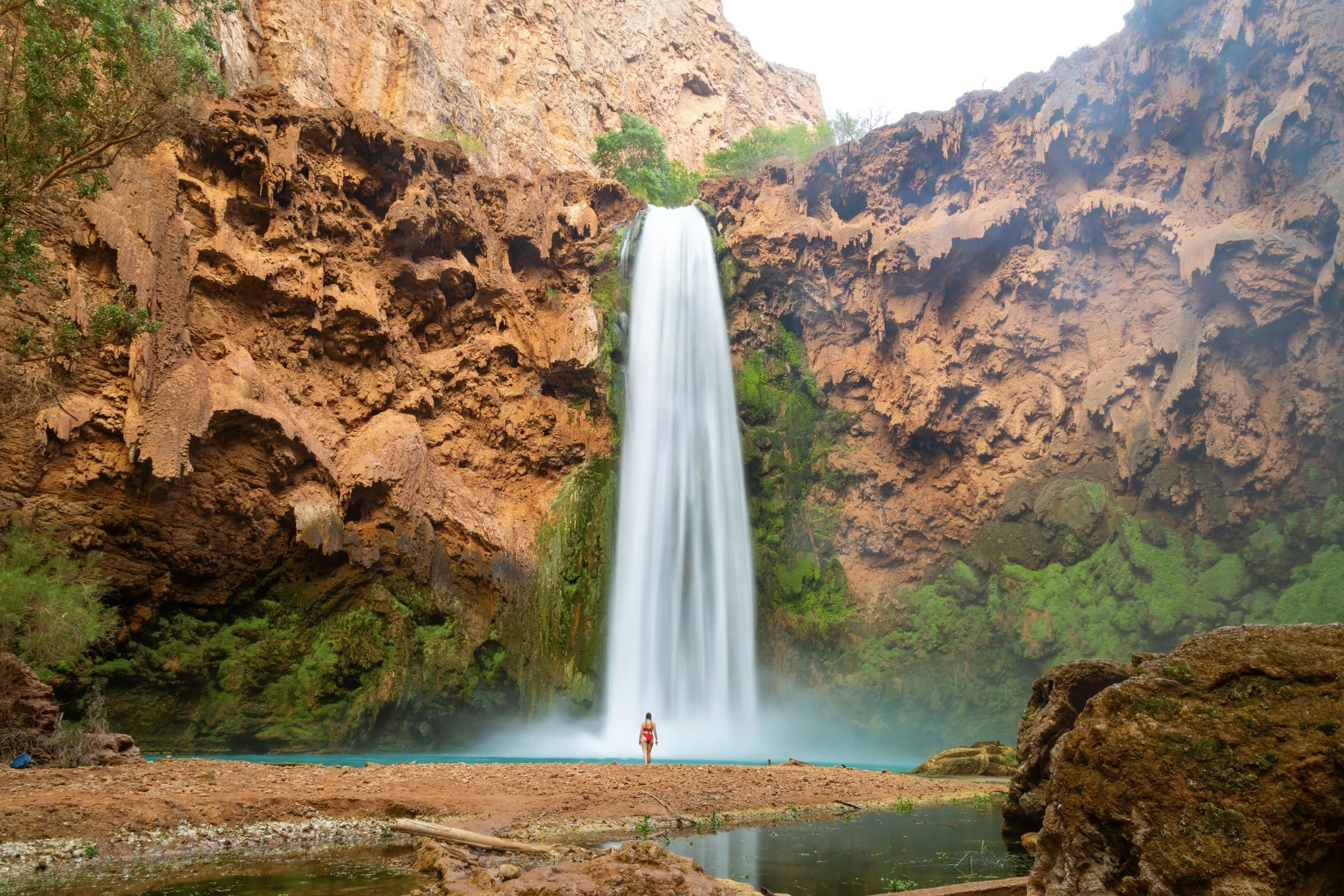 A woman stands in awe beneath the towering Mooney Falls waterfall, surrounded by the red rock canyon walls and lush moss-covered cliffs of Havasupai, Arizona.