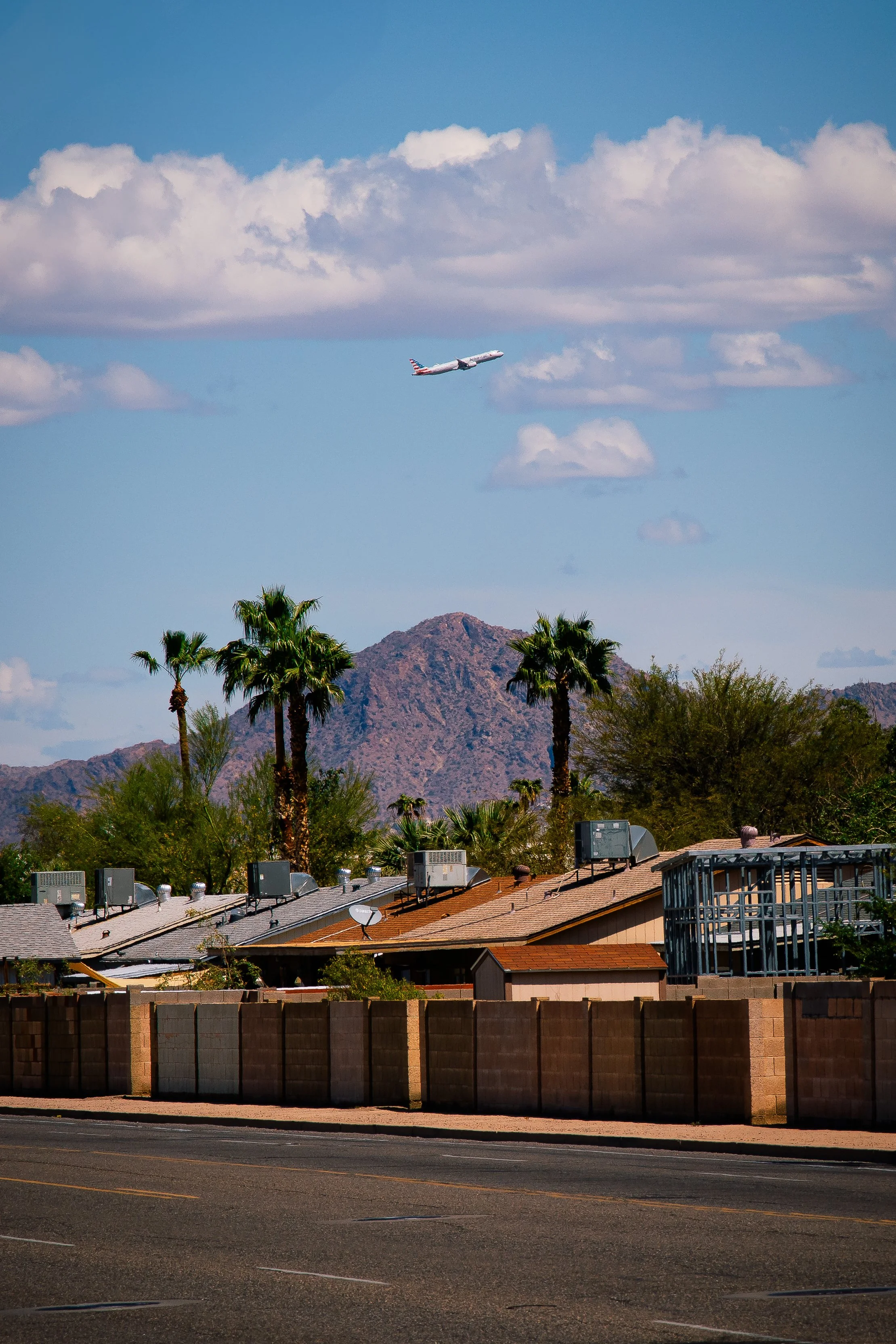 A distant airplane soars above a Phoenix neighborhood with Camelback Mountain in the background and palm trees in the foreground under a partly cloudy sky.