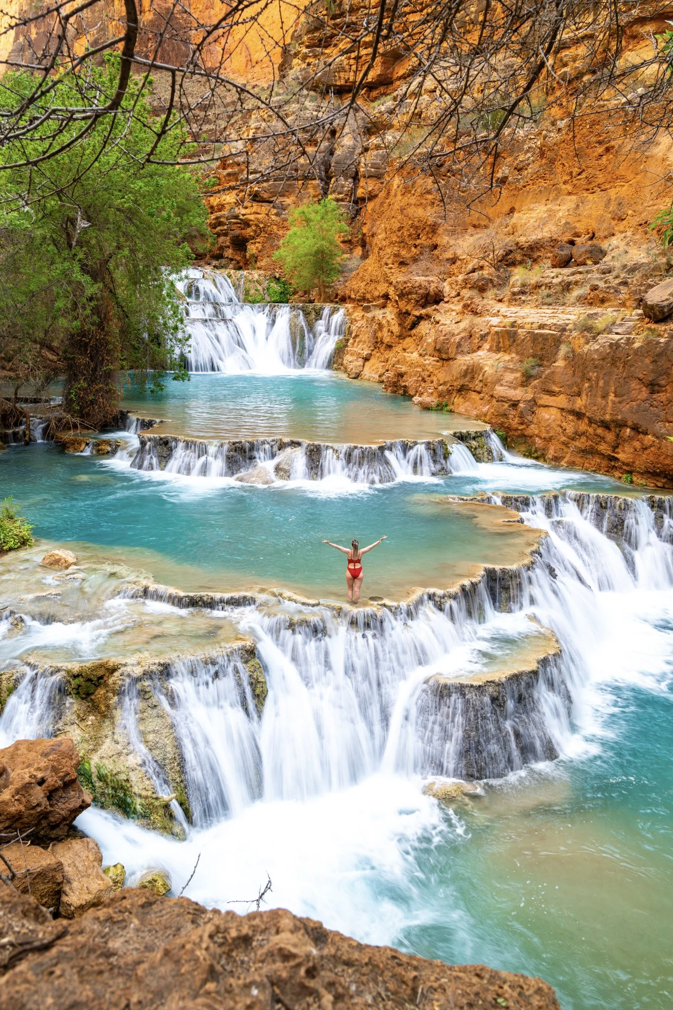 A woman stands with arms raised in a red swimsuit on a limestone ledge between cascading turquoise waterfalls at Beaver Falls in Havasupai, surrounded by red rock canyon walls and desert foliage.
