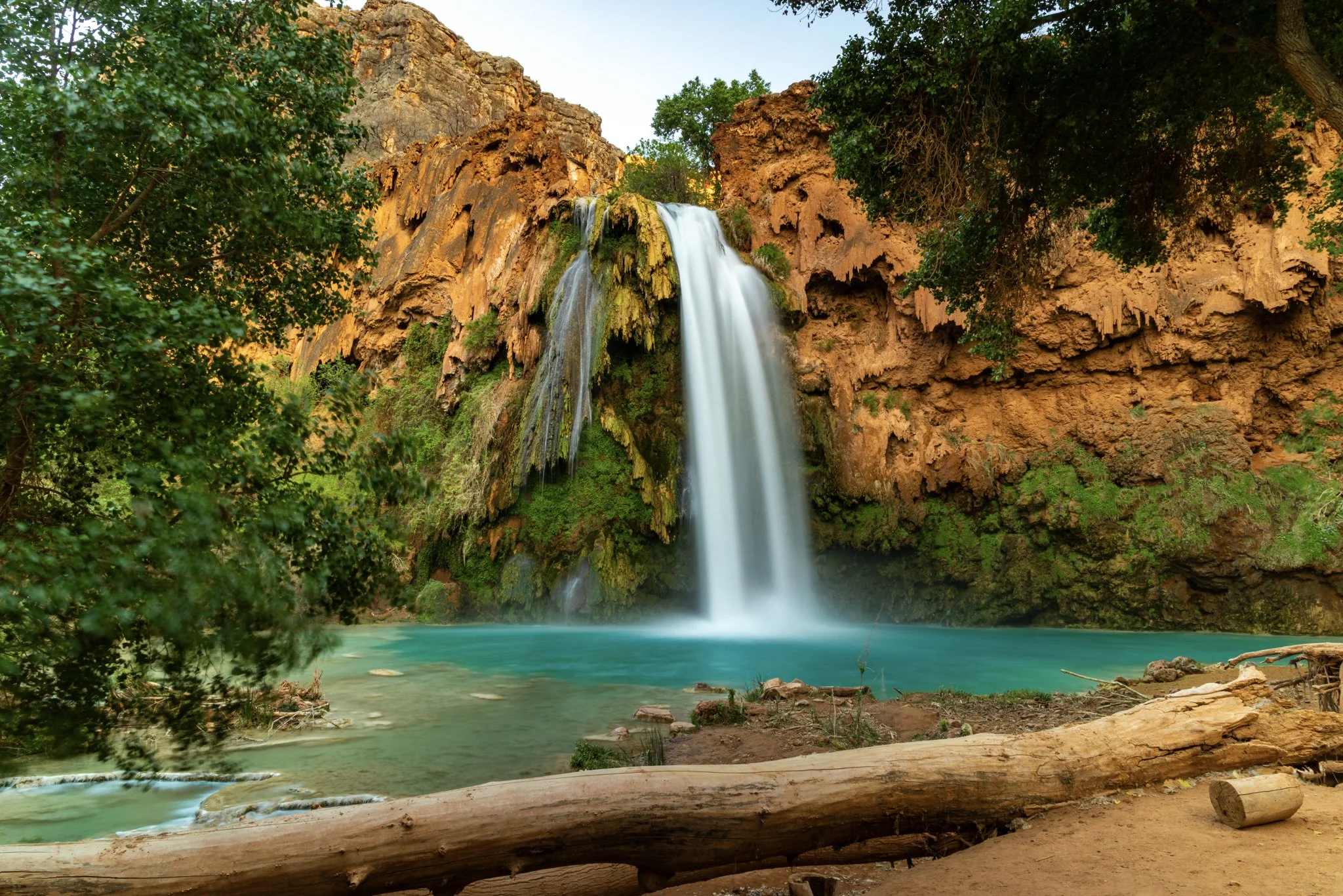 A wide-angle photo of Havasu Falls cascading into a turquoise pool, surrounded by red canyon walls and green vegetation at sunrise in Havasupai Canyon, Arizona.