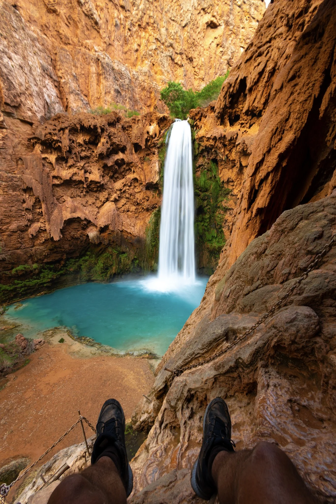 Point-of-view image of a hiker’s legs over a cliff ledge overlooking Mooney Falls, a tall waterfall plunging into a turquoise pool framed by red canyon walls and greenery in Havasupai, Arizona.