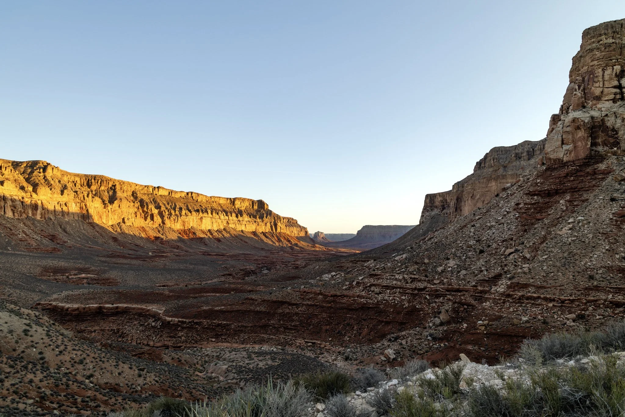Early morning sunlight striking the canyon walls along the Hualapai Trailhead, with a vast desert landscape leading into the Grand Canyon’s red rock formations under a clear sky.