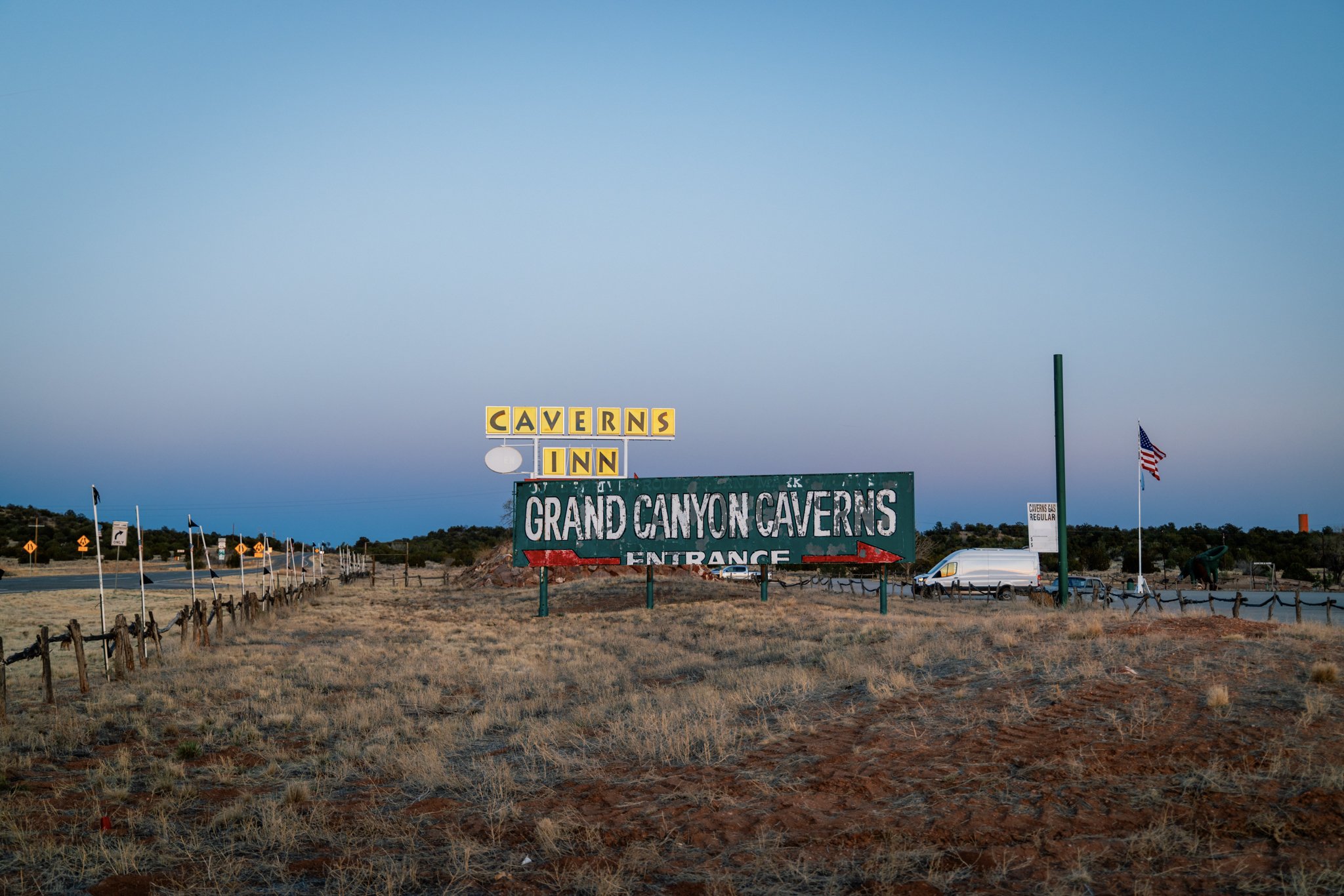A weathered green sign reading “Grand Canyon Caverns Entrance” stands beneath a twilight sky near the check-in area for Havasupai permit holders, with a white van and an American flag visible in the background