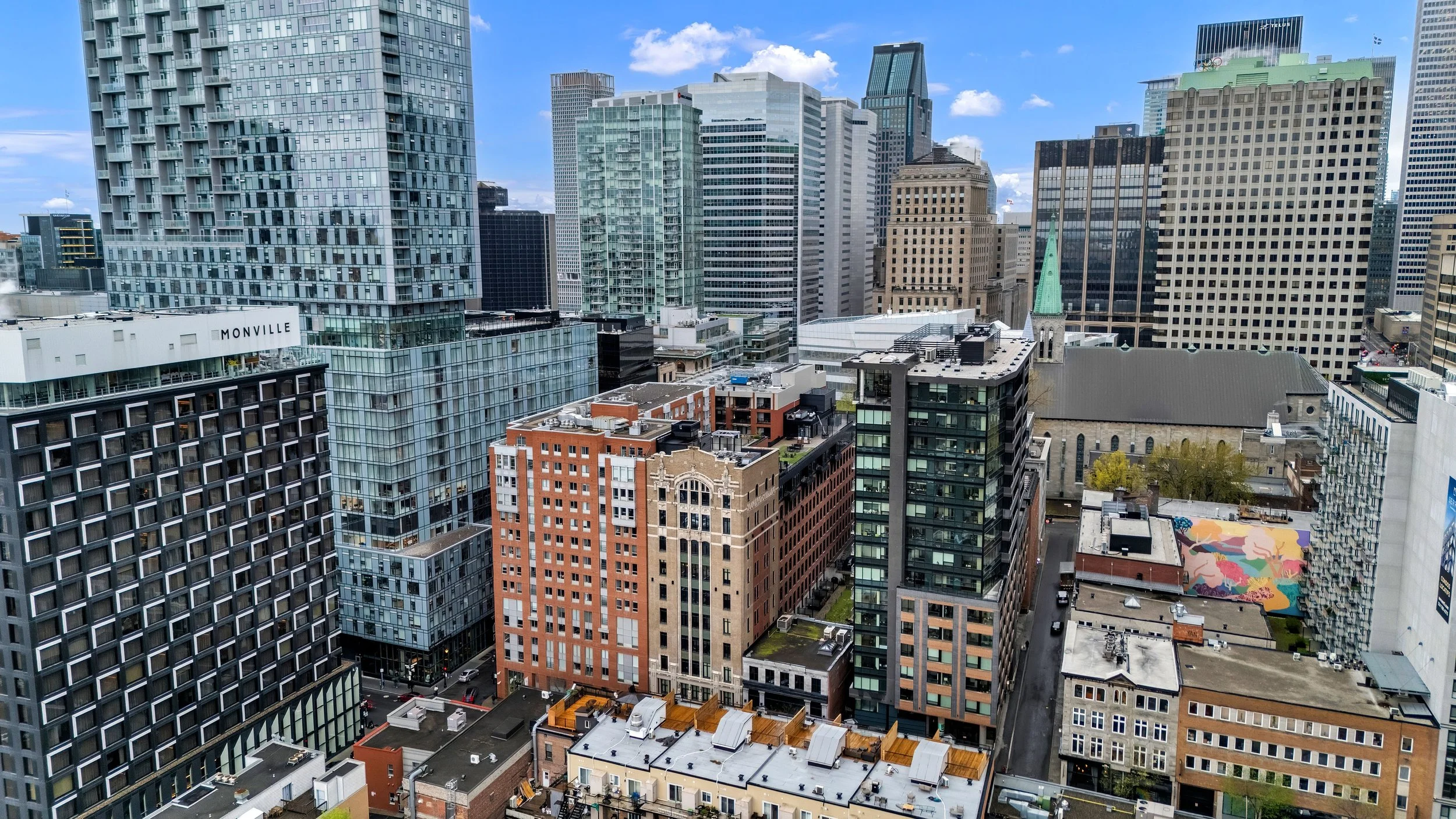 A city skyline with numerous modern high-rise buildings, including glass skyscrapers, traditional brick structures, and a church with a green steeple under a partly cloudy sky.