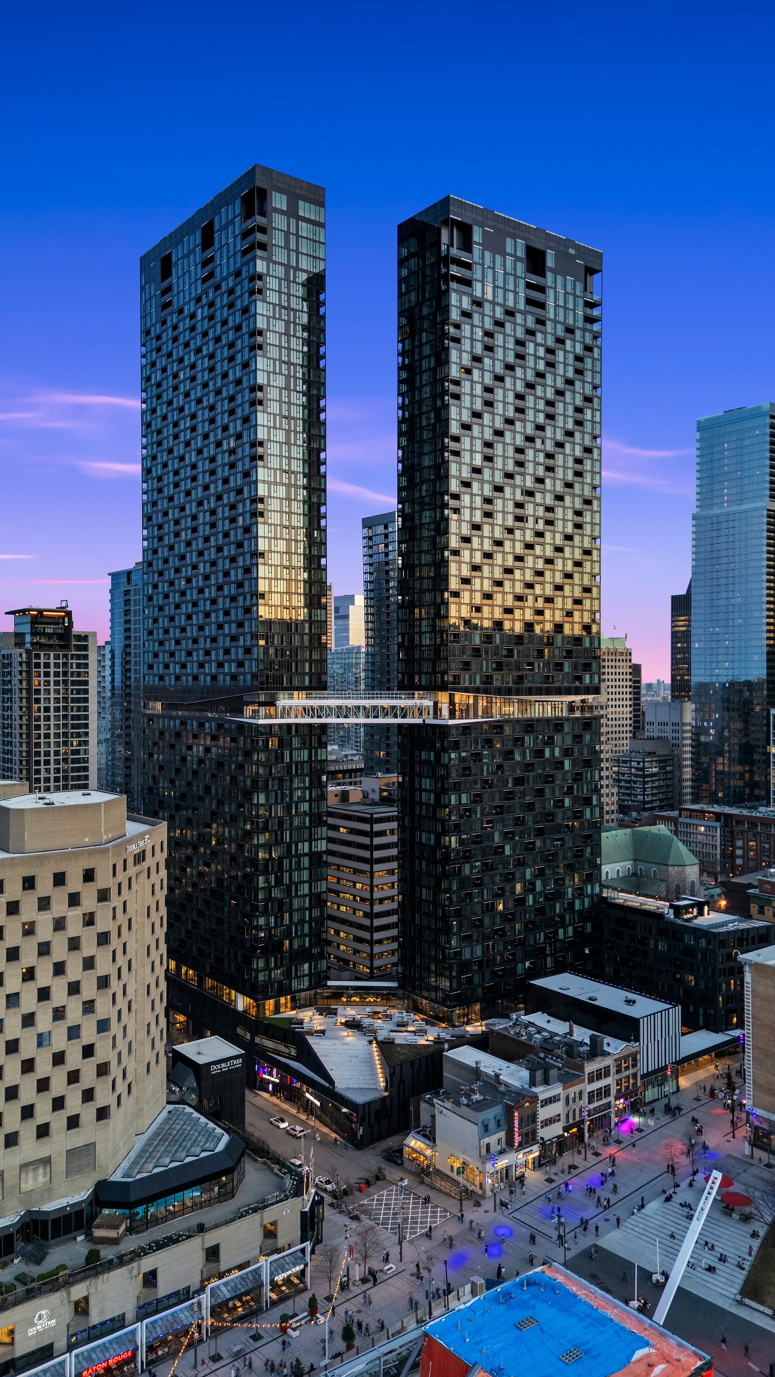 Tall modern skyscrapers in a city at dusk with a busy outdoor plaza below filled with people, lights, and ice skating rink.