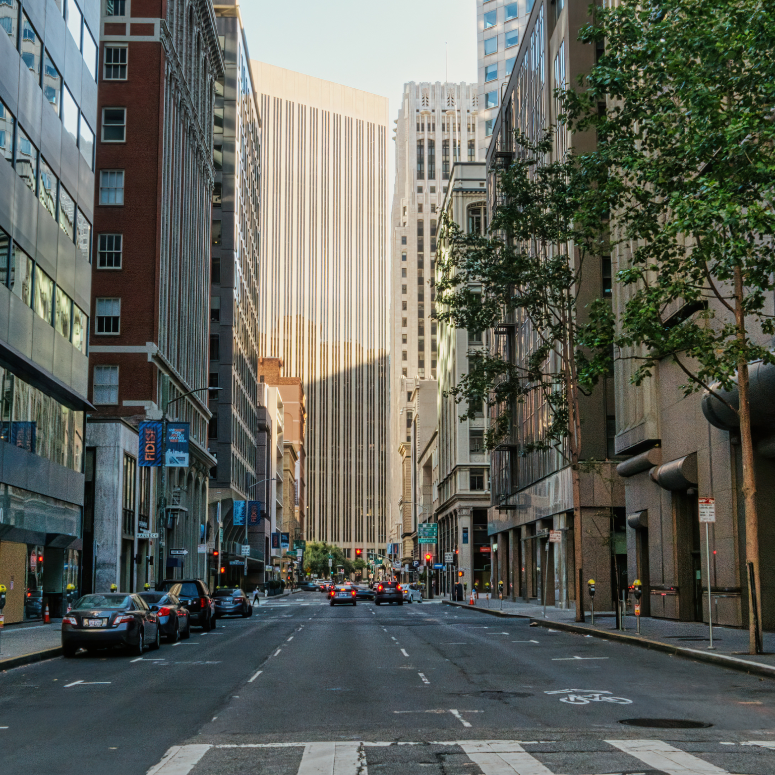 City street view with tall buildings, cars parked along the sides, and a few cars driving down the road. Trees line the sidewalks, and there are traffic lights at the intersection.