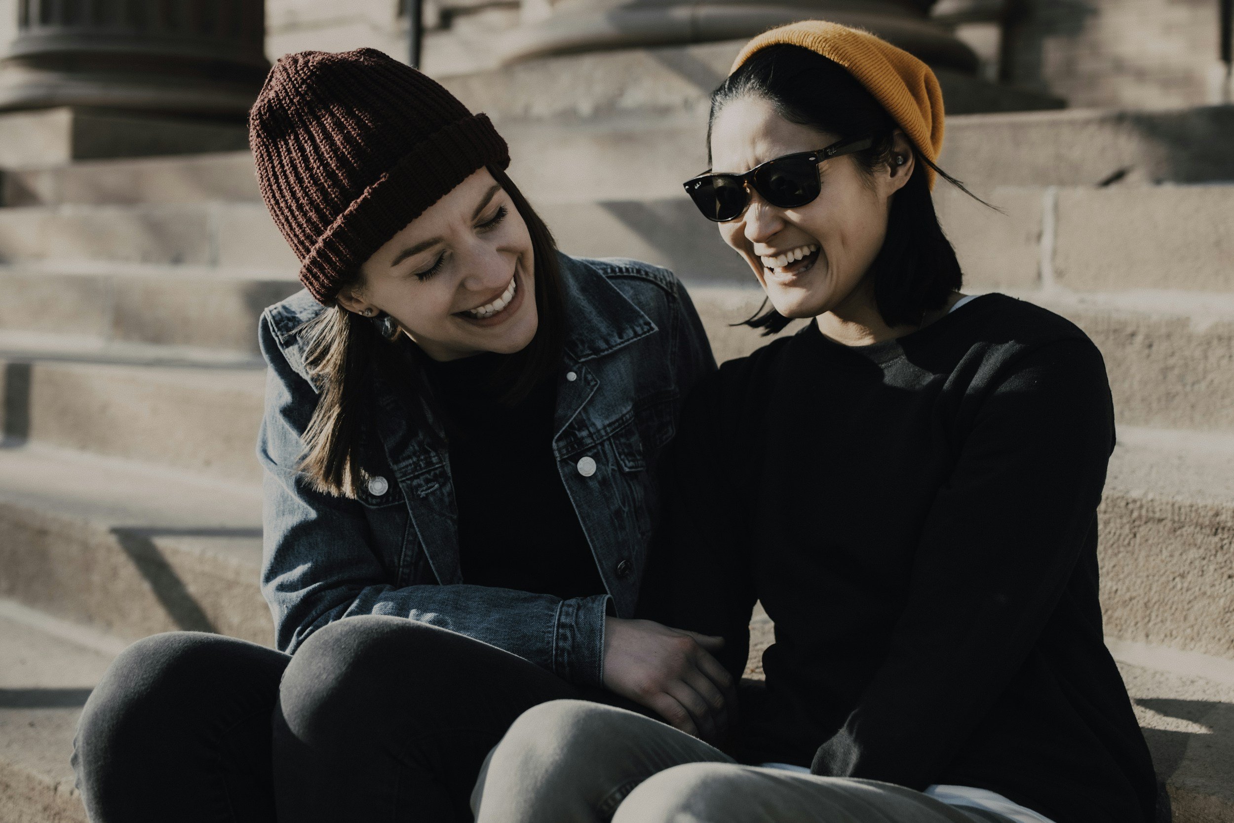 Two young women sit on outdoor steps, smiling and laughing together during daytime. One wears a burgundy knit hat and denim jacket, the other sunglasses and an orange beanie.