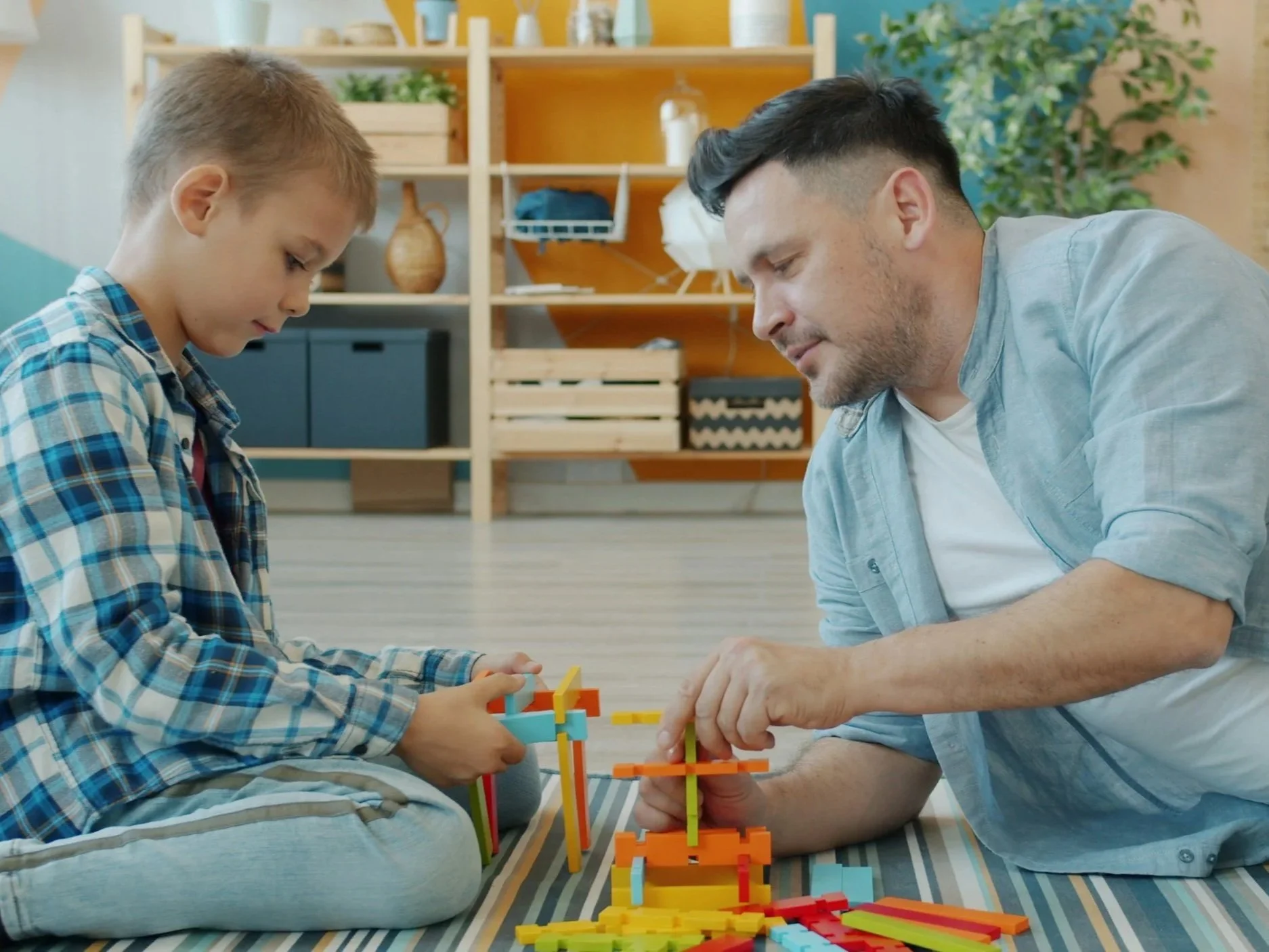 A young boy with a plaid shirt and an adult man with a light blue shirt are sitting on a striped rug, playing with colorful building blocks, building a structure together. They are in a cozy room with wooden shelves, decorative items, and green plants in the background.