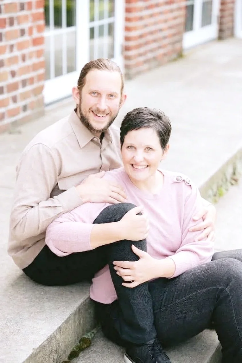 A man and a woman sitting on a sidewalk outside a brick building, smiling at the camera. The man has long hair and a beard, wearing a beige shirt, and the woman has short dark hair, wearing a pink sweater. The woman is holding a black dog wrapped in a pink blanket.