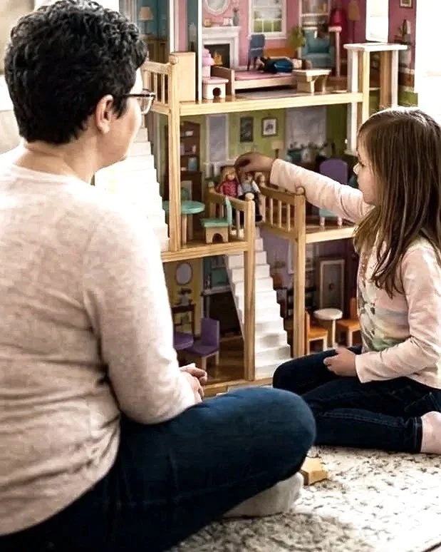 A girl and an adult sitting on the floor in a playroom, playing with a large dollhouse.