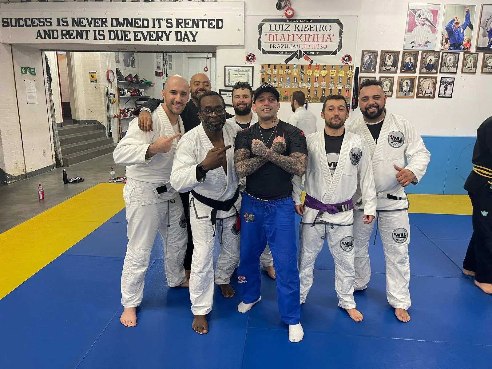 Group of men in a Brazilian Jiu-Jitsu dojo, wearing gi uniforms, posing together on a blue mat. A motivational quote is on the wall behind them.