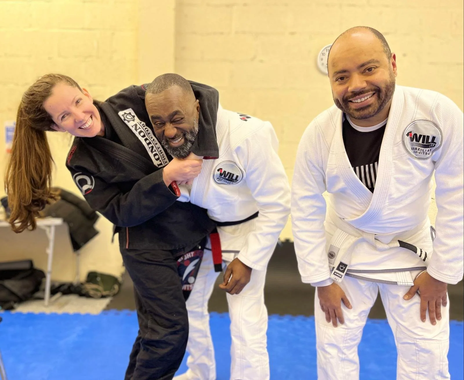 Three people in Brazilian jiu-jitsu uniforms smiling, with one person playfully holding another in a headlock.