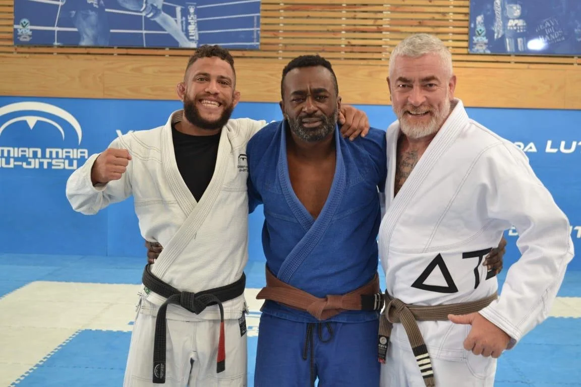 Three men wearing martial arts gi, posing together in a gym with "Damian Maia Jiu-Jitsu" signage in the background.