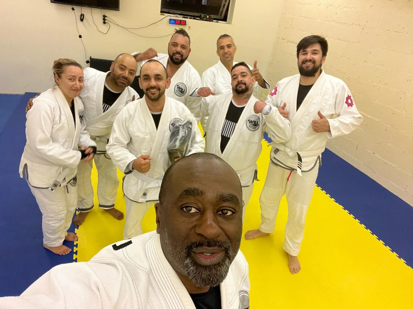 Group of people wearing white martial arts uniforms, standing on a blue and yellow mat, posing for a selfie in a gym or dojo.