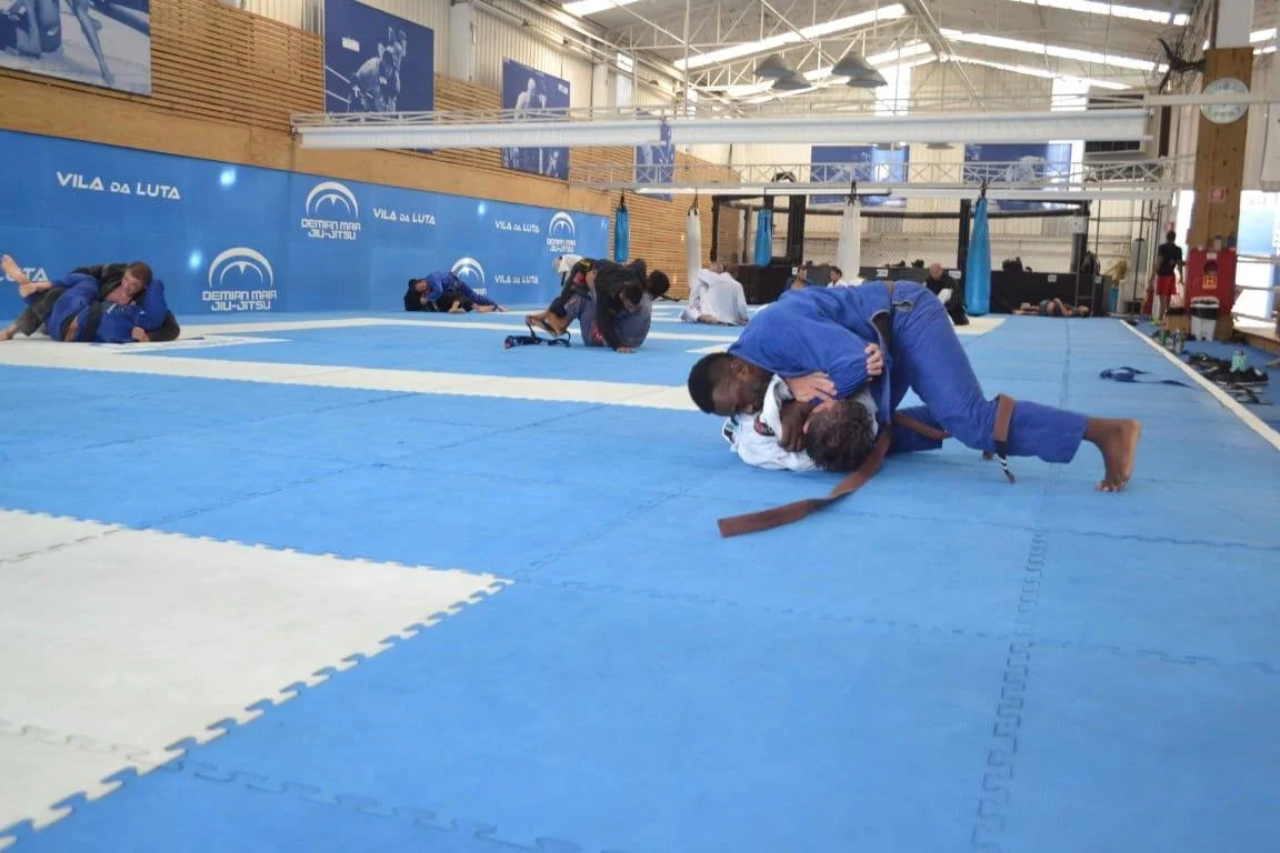 People practicing Brazilian Jiu-Jitsu in a training facility with blue mats and branded walls.