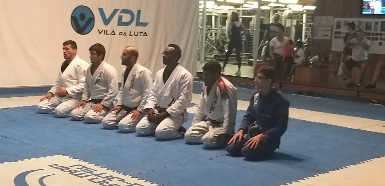 Group of people in martial arts uniforms kneeling on a blue mat in a gym.
