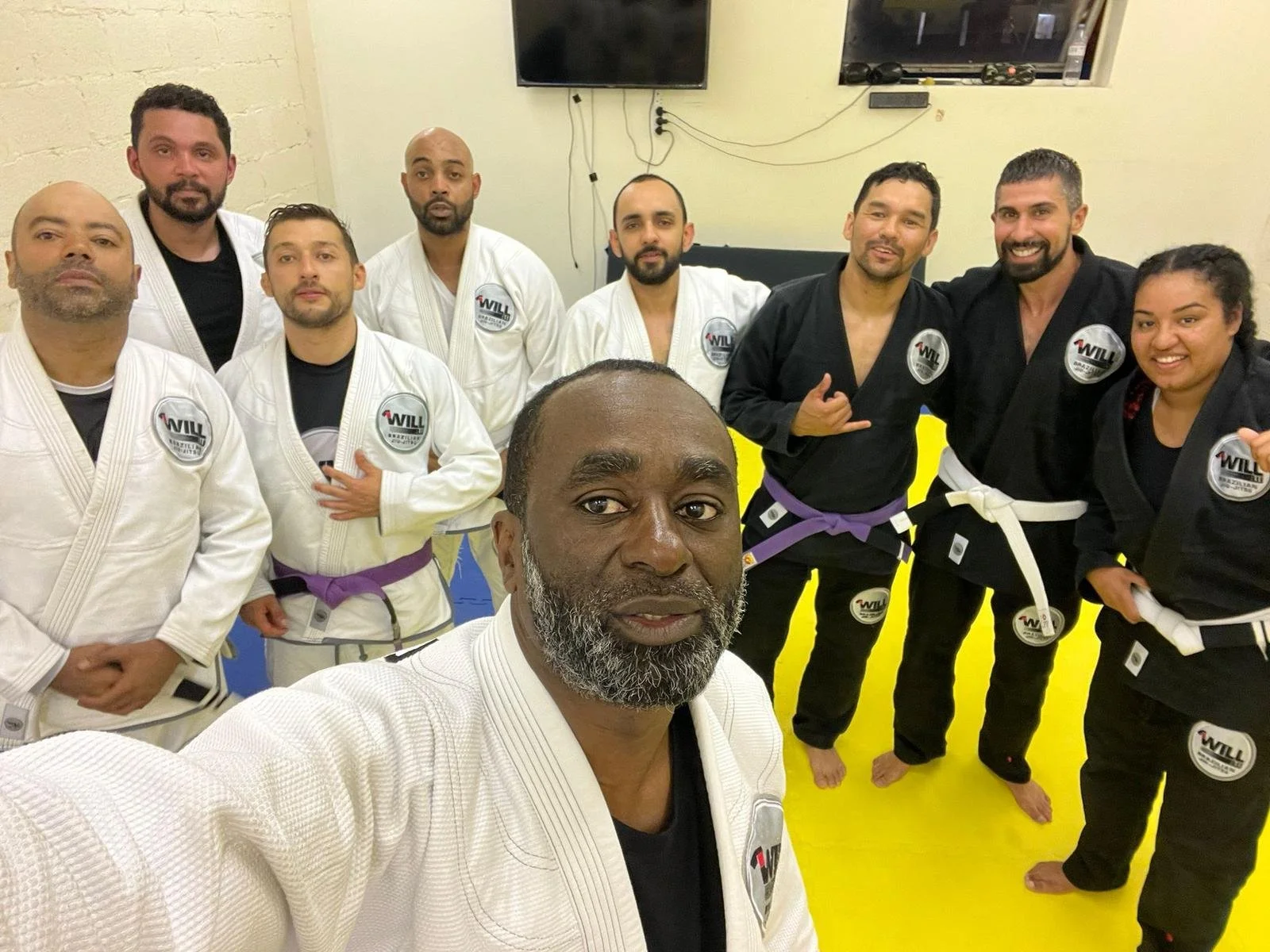 Group of people in martial arts uniforms posing in a training room with a yellow mat and TV screens on the wall.