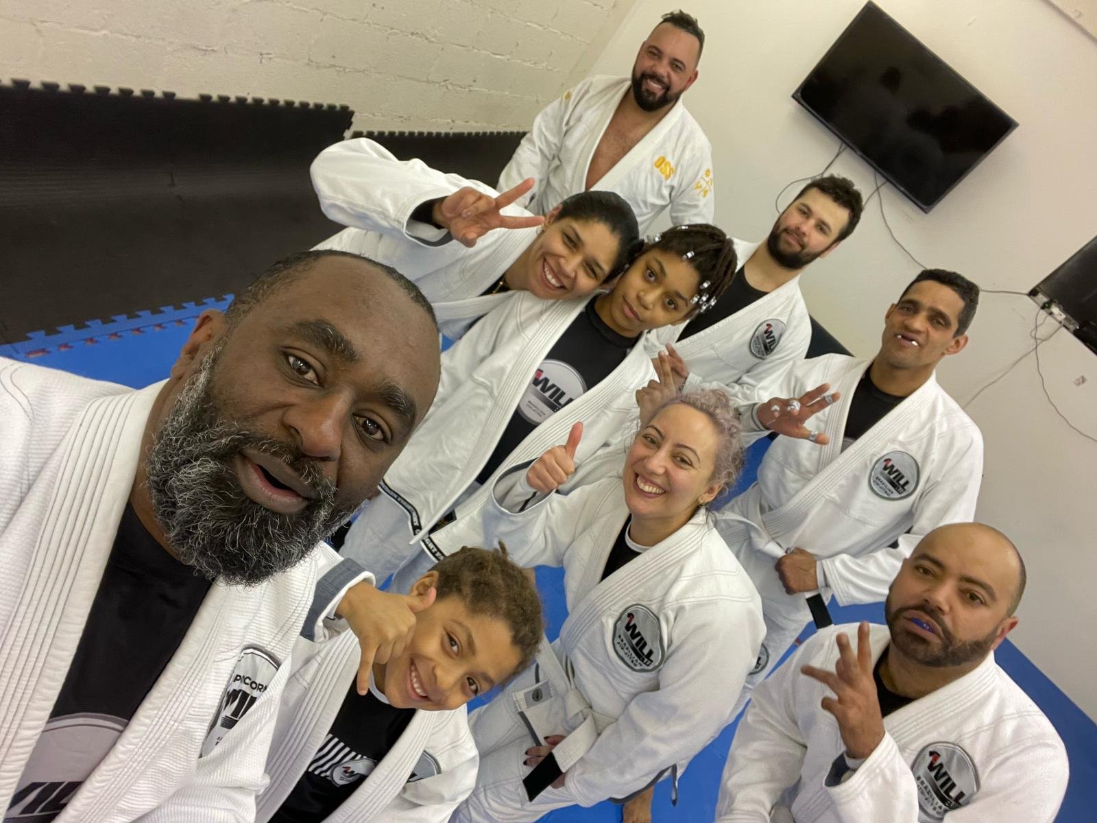 Group of people in martial arts uniforms posing in a dojo, smiling and making peace signs.