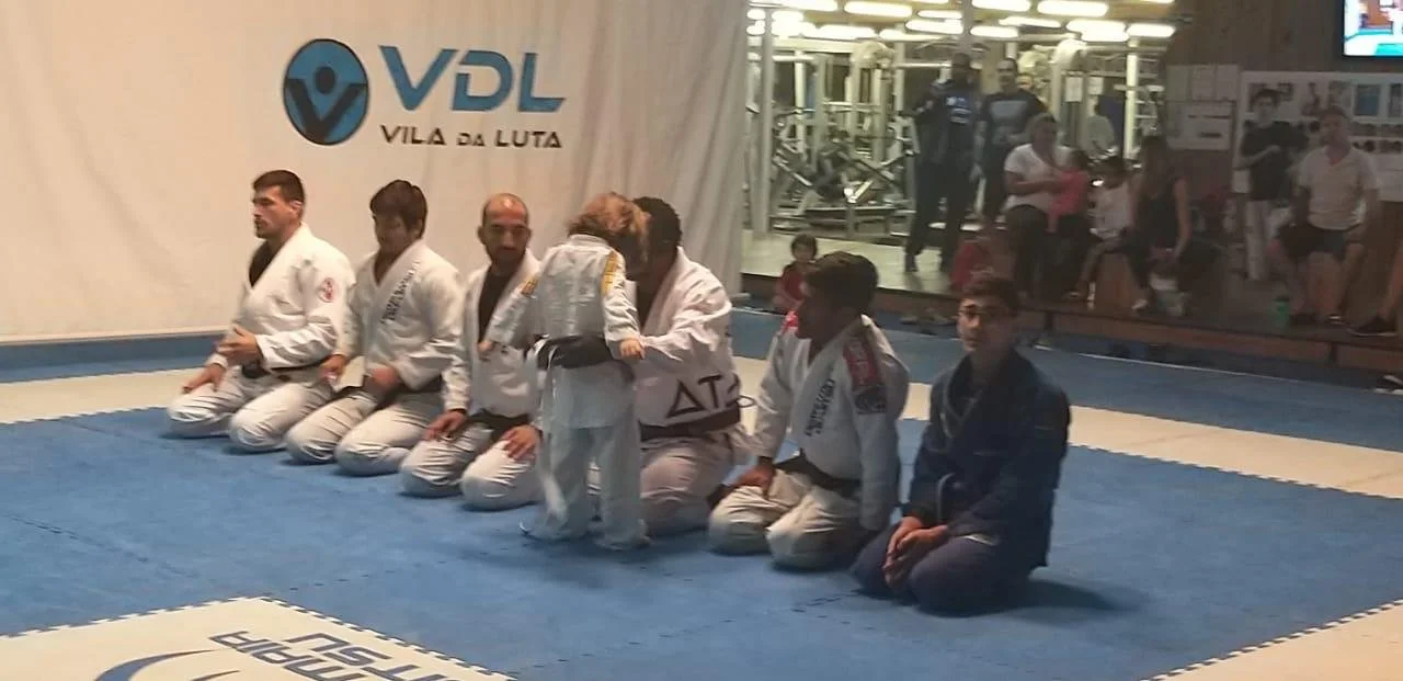 A group of people practicing martial arts in a gym, with several participants wearing white and blue judo gis kneeling in a line, including a small child being assisted by an adult. The gym has a banner reading 'Vila da Luta' in the background and people sitting on benches observing.