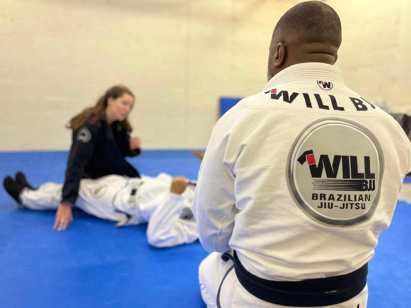 People in Brazilian Jiu-Jitsu gis practicing on a blue mat, focusing on one person with a black belt and "Will BJJ" on the back of their gi.