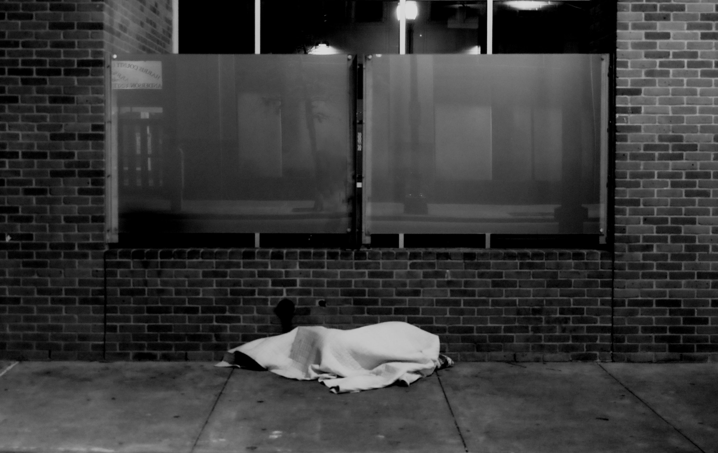 Nighttime street scene showing a wrapped body on the sidewalk in front of a brick building with a metal gate and large windows.