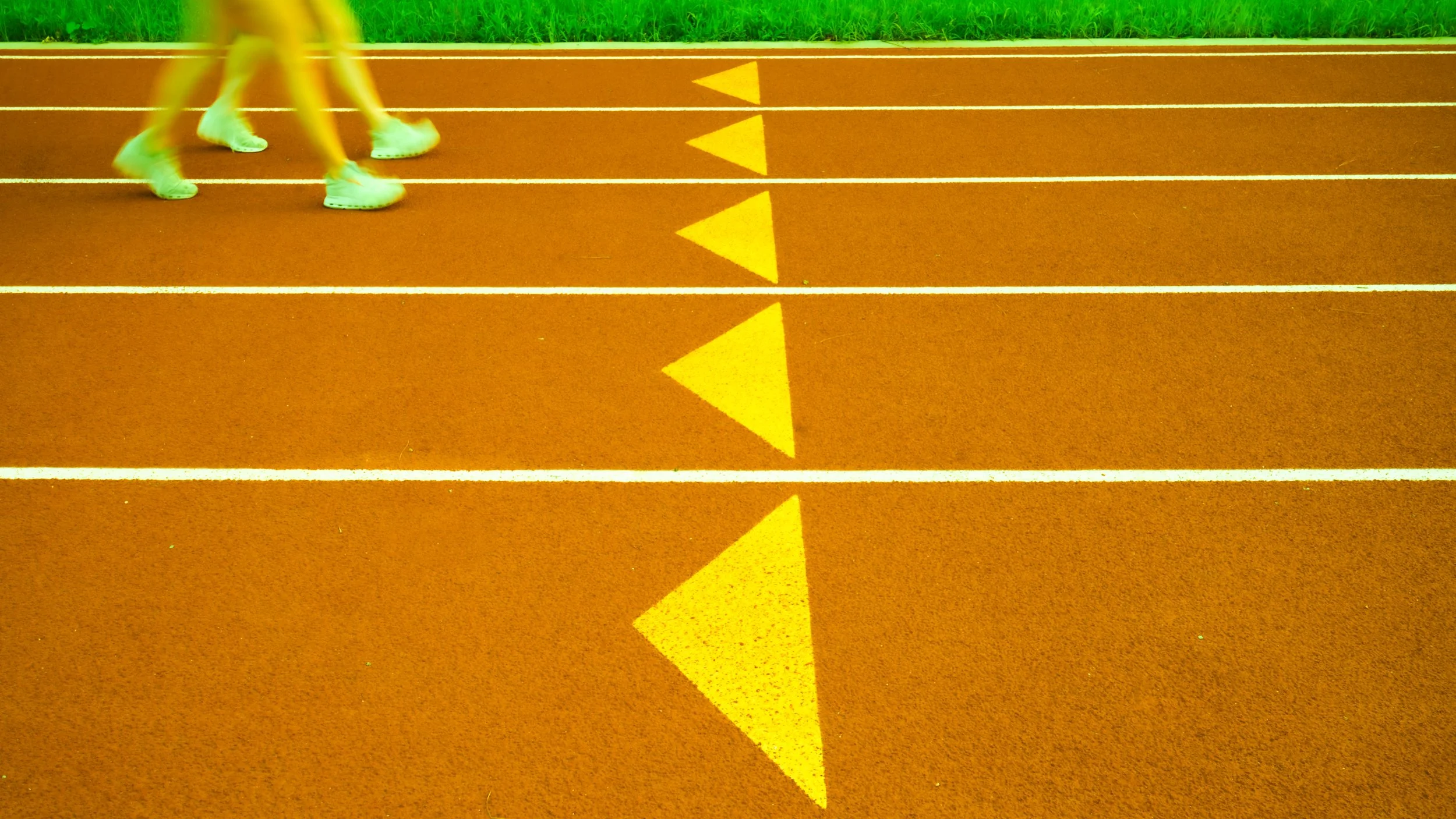 A running track with yellow directional triangles on the ground. A person's legs and feet are visible, wearing white running shoes and shorts, mid-stride on the track.