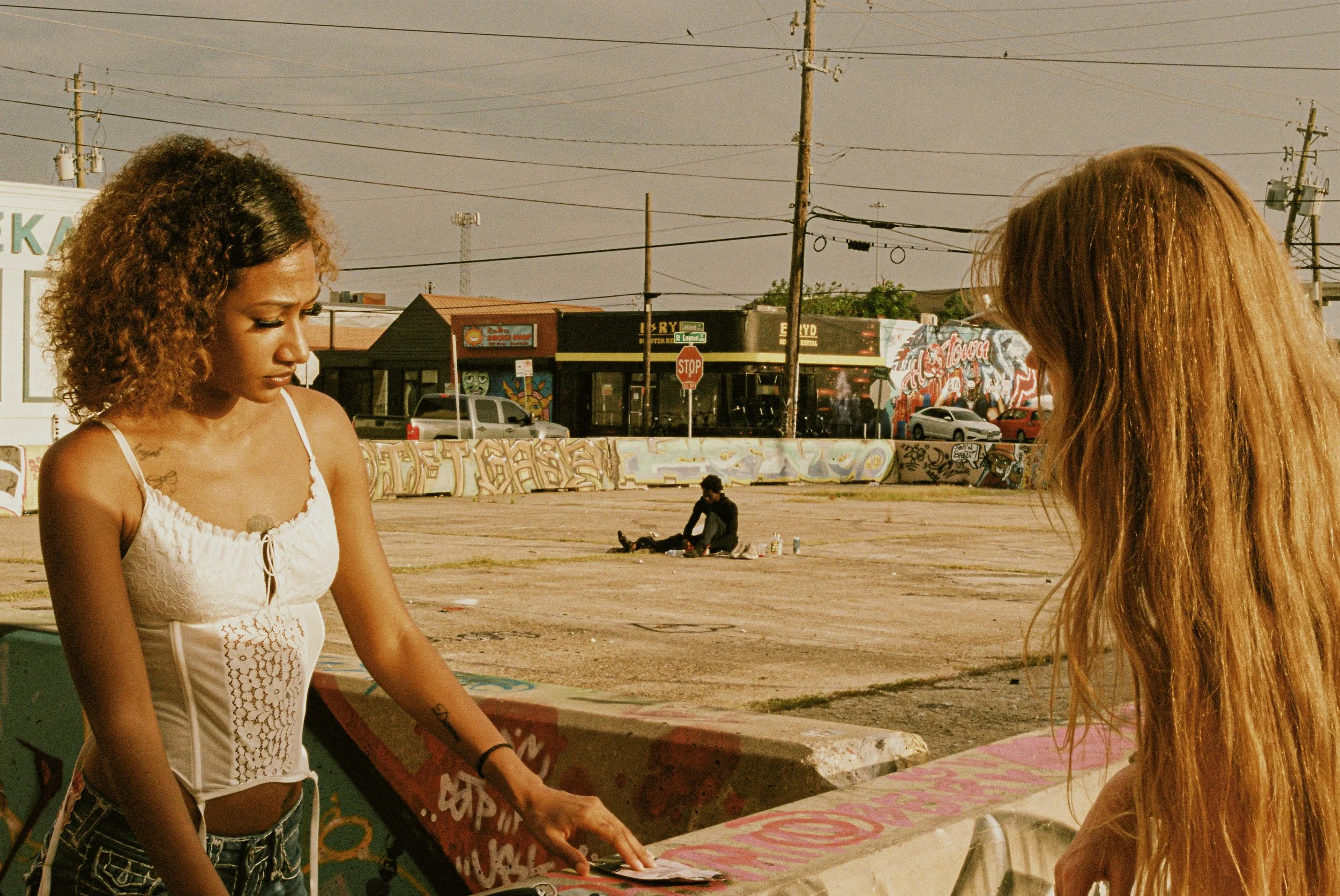 Two young women with long hair standing at a graffiti-covered counter outdoors, with a person sitting on the ground in the background, against a backdrop of graffiti walls, utility poles, and buildings.