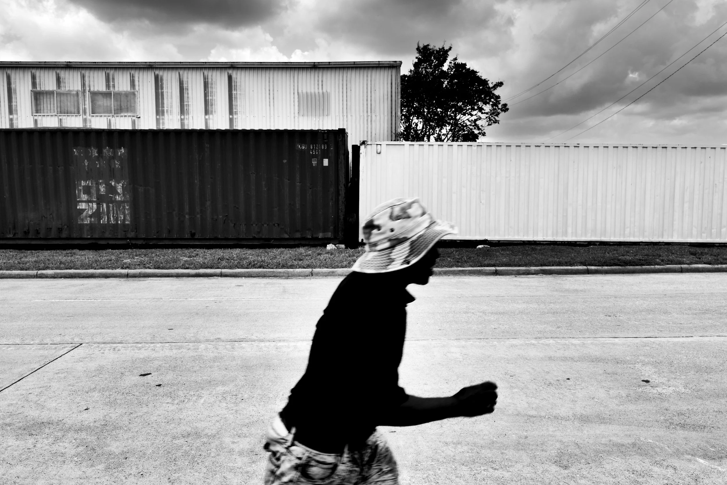 A person wearing a bucket hat and shorts walking along a sidewalk with a background of metal storage containers and a building, in black and white.
