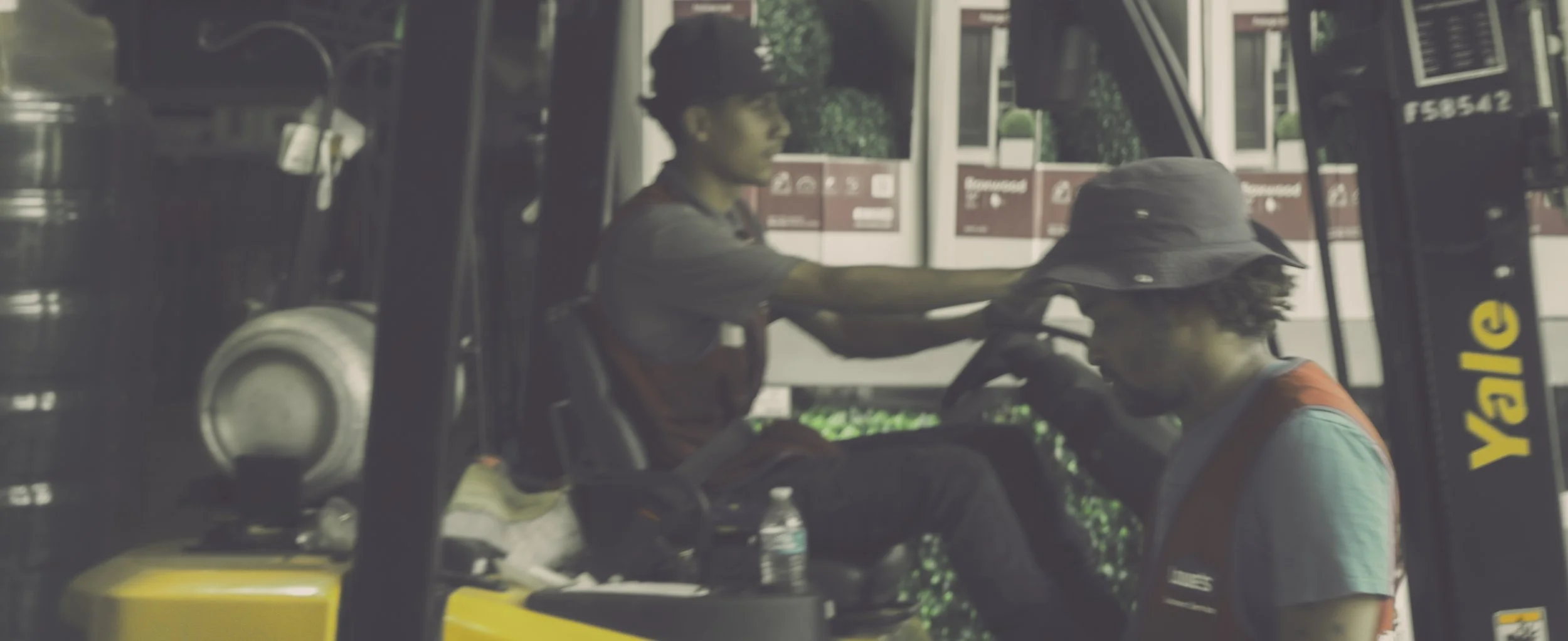 A person sitting in a yellow forklift, another person leaning on the forklift's seat, both wearing casual clothes and hats, with a gas station in the background.
