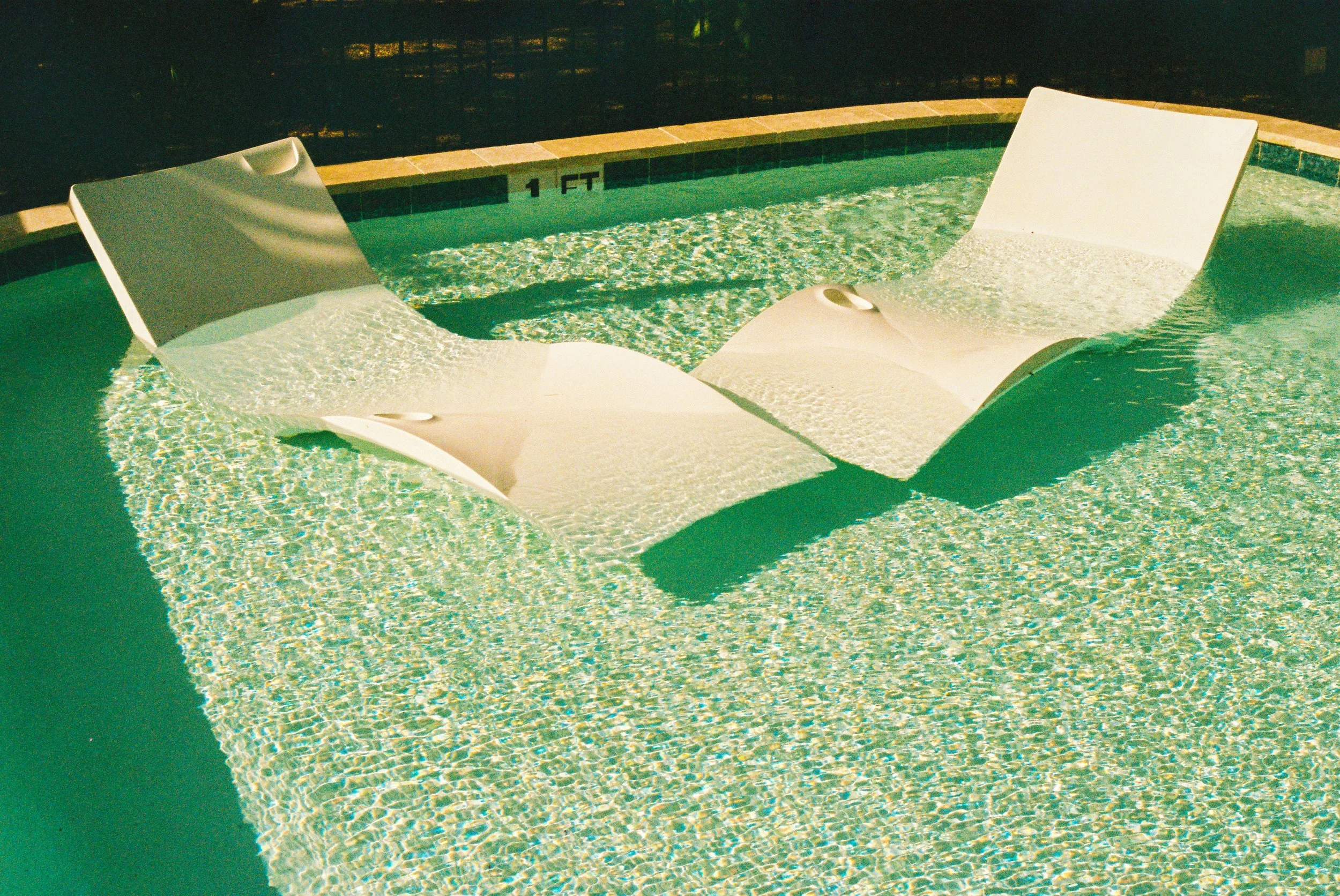 Two white pool loungers submerged in a swimming pool with water ripples, near the 1 foot depth marker.