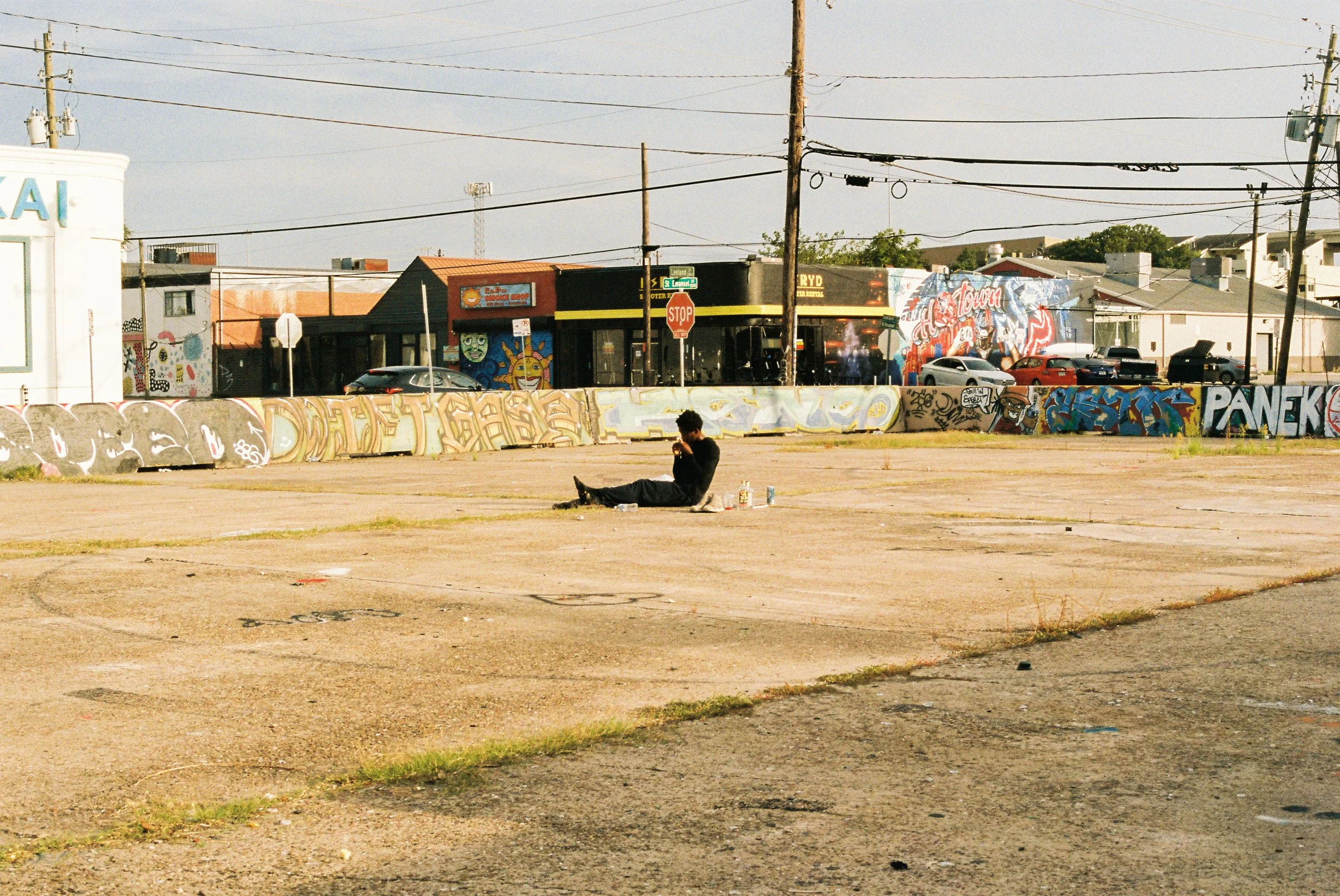 A person sitting on the ground in an empty lot, drinking from a cup or bottle, with graffiti on a wall and buildings with parked cars in the background.