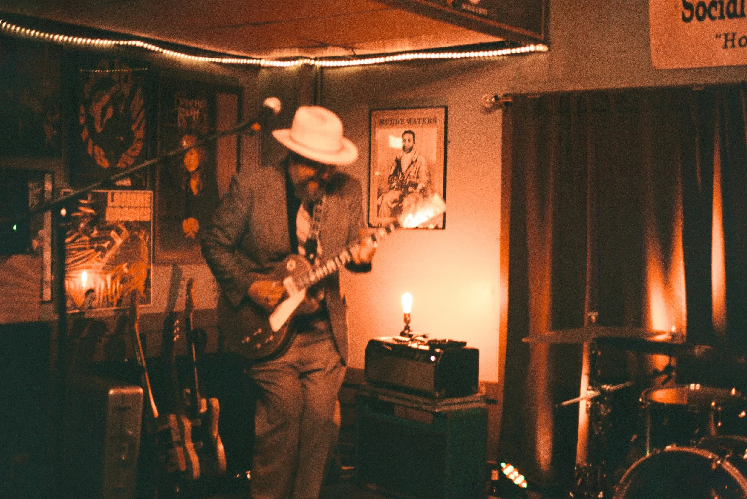 A man wearing a hat, glasses, and a suit jacket playing an electric guitar on stage in a dimly lit room decorated with posters and artwork.